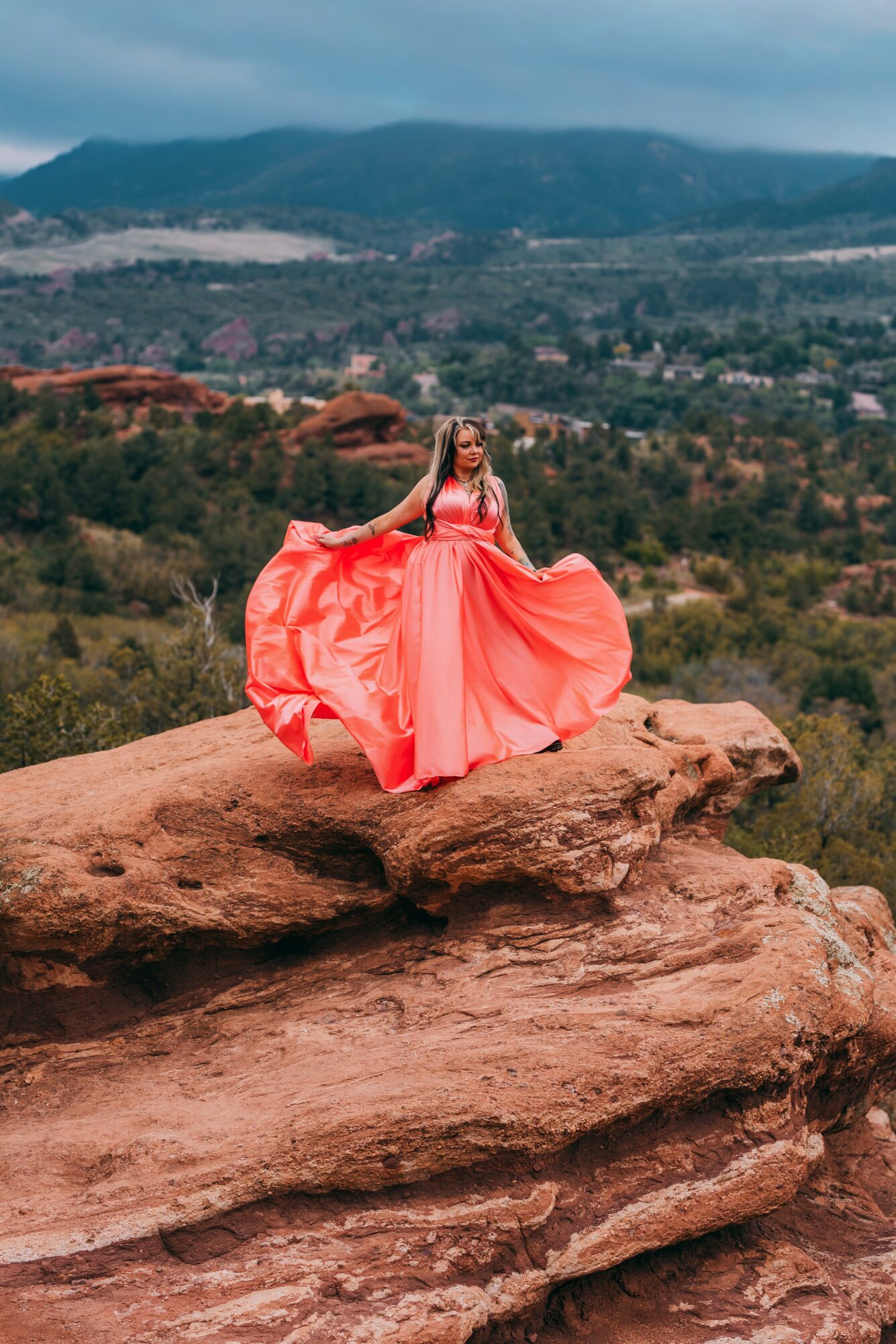 Woman in a flowing pink dress standing on a large rock with a landscape of hills and trees in the background.