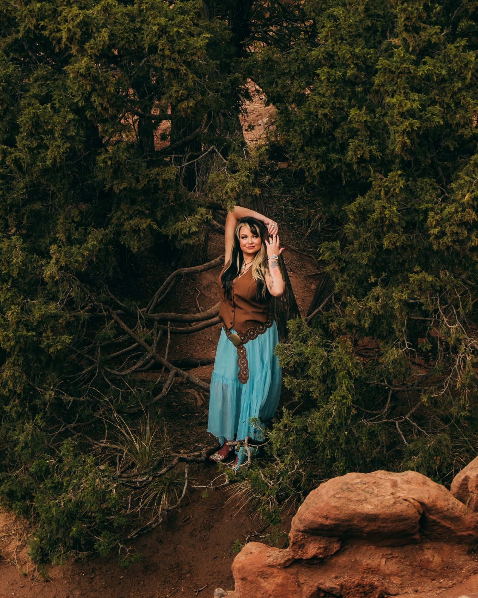 Woman standing among trees and rocks, wearing a brown top and blue skirt, with one hand on her head.