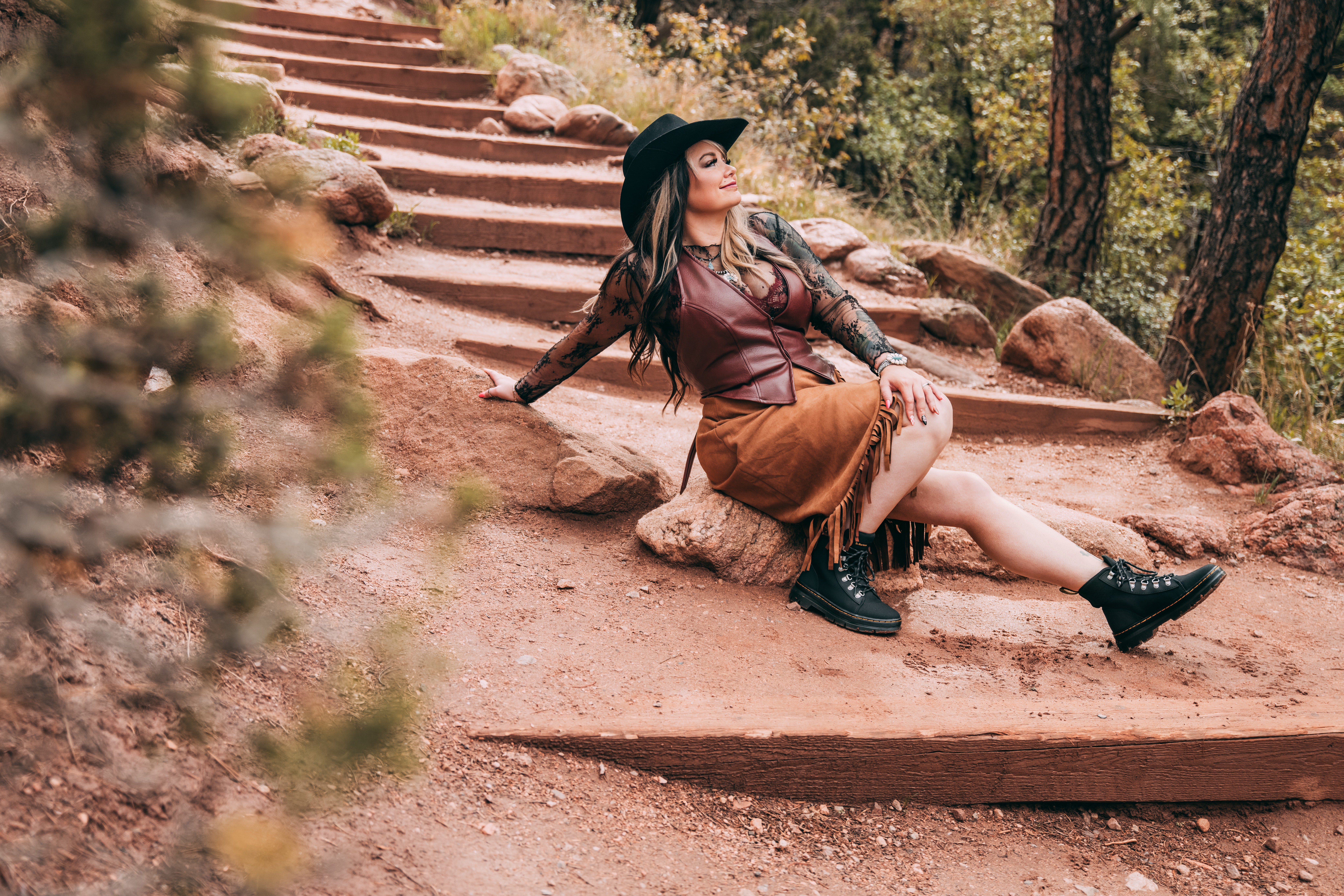Woman sitting on a rock on a dirt trail with steps, surrounded by trees and foliage, wearing a hat and brown skirt.