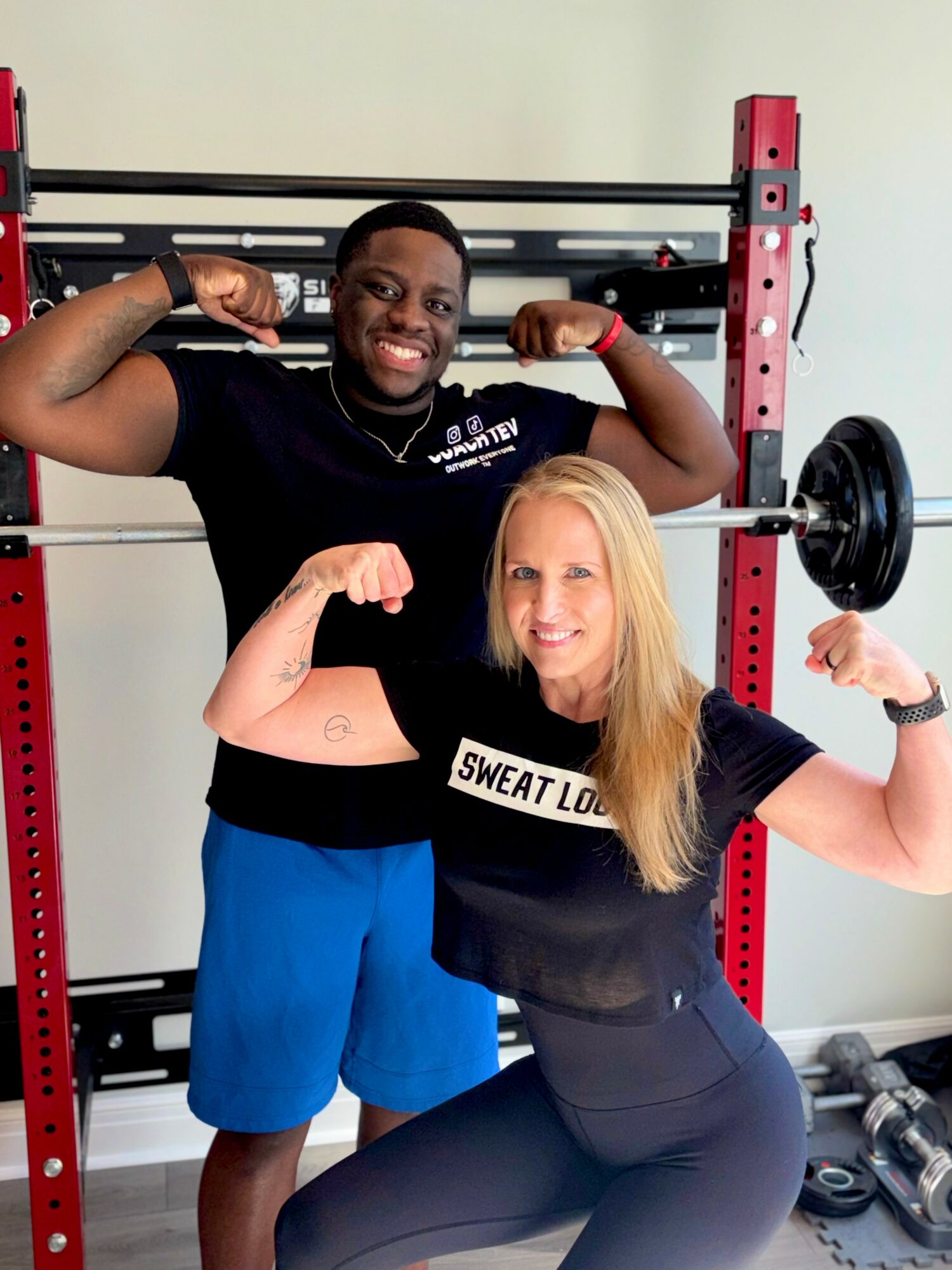 Two people flexing their arm muscles in a gym, smiling, with weightlifting equipment in the background.
