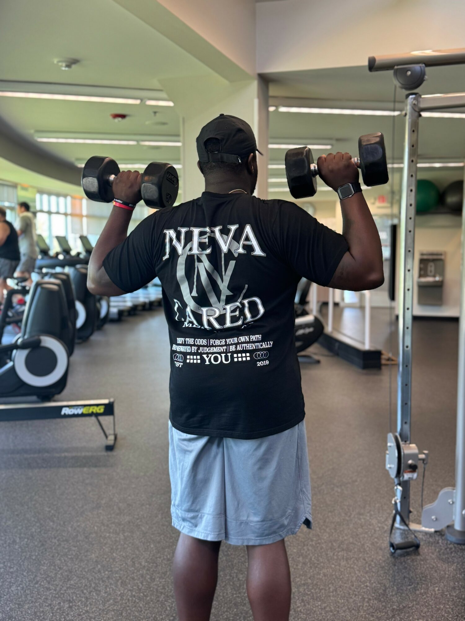 Person lifting dumbbells in a gym, facing away, wearing a black cap and T-shirt, with gym equipment in background.