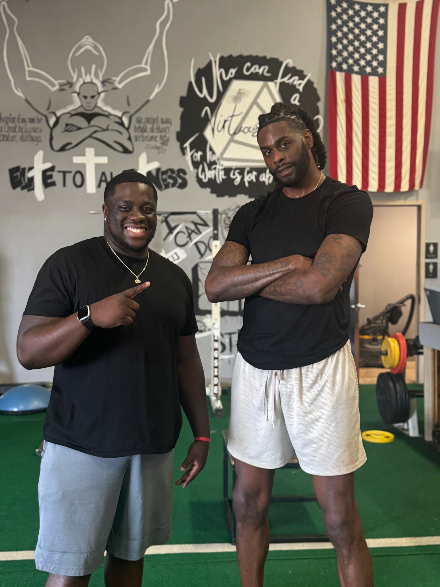 Two men standing in a gym, one smiling and pointing, the other with arms crossed, gym equipment in background.