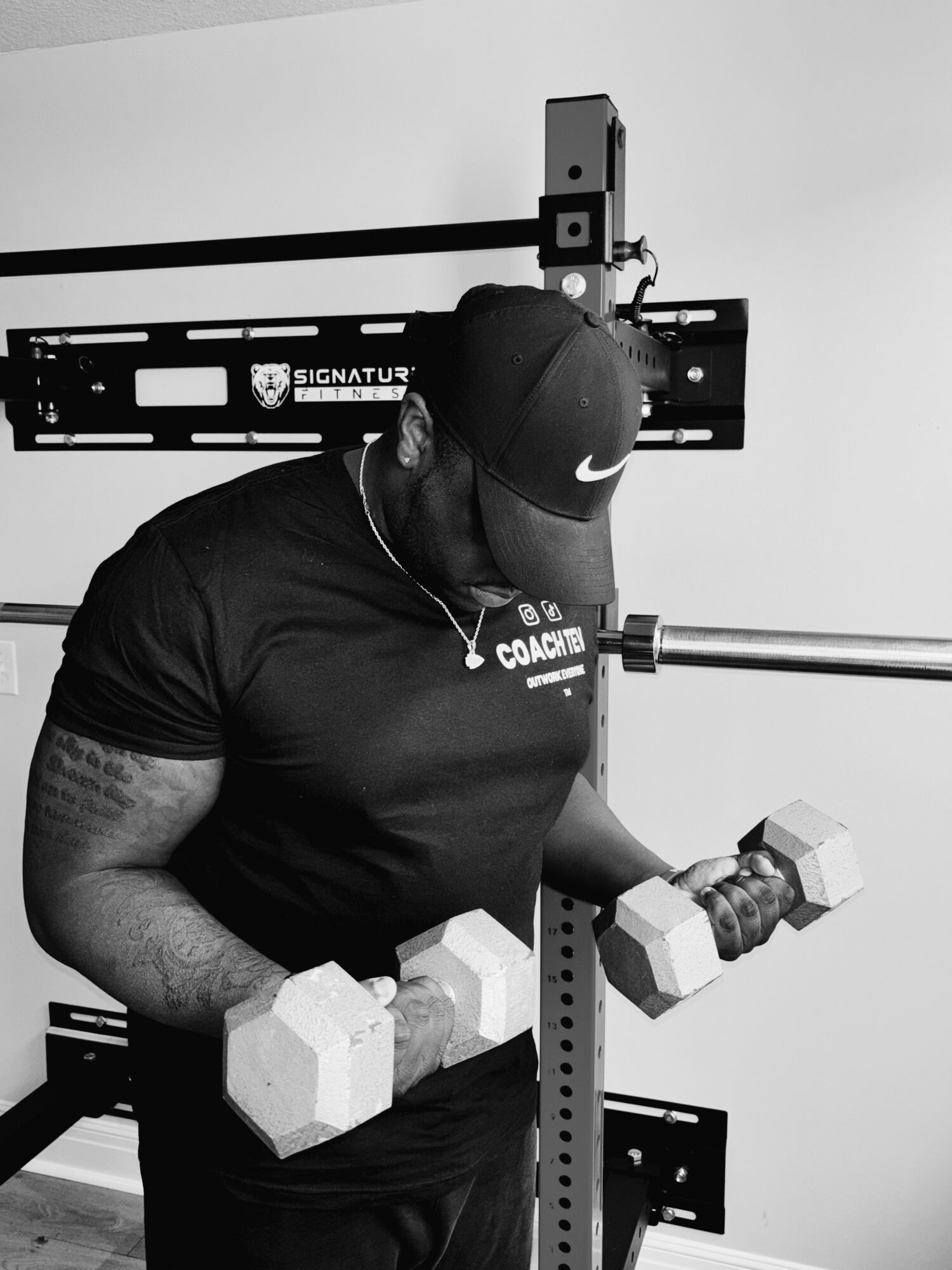 Man in black shirt and cap lifting dumbbells in a gym, with workout equipment in background.