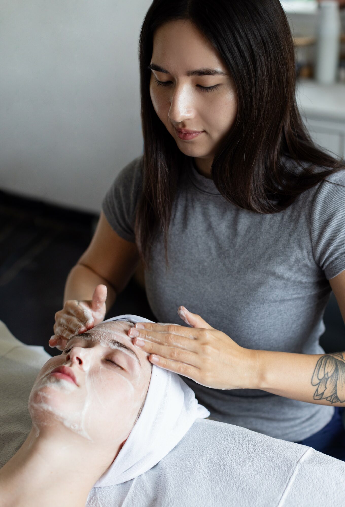 A woman applies a facial mask to a person lying down with a towel wrapped around their head.