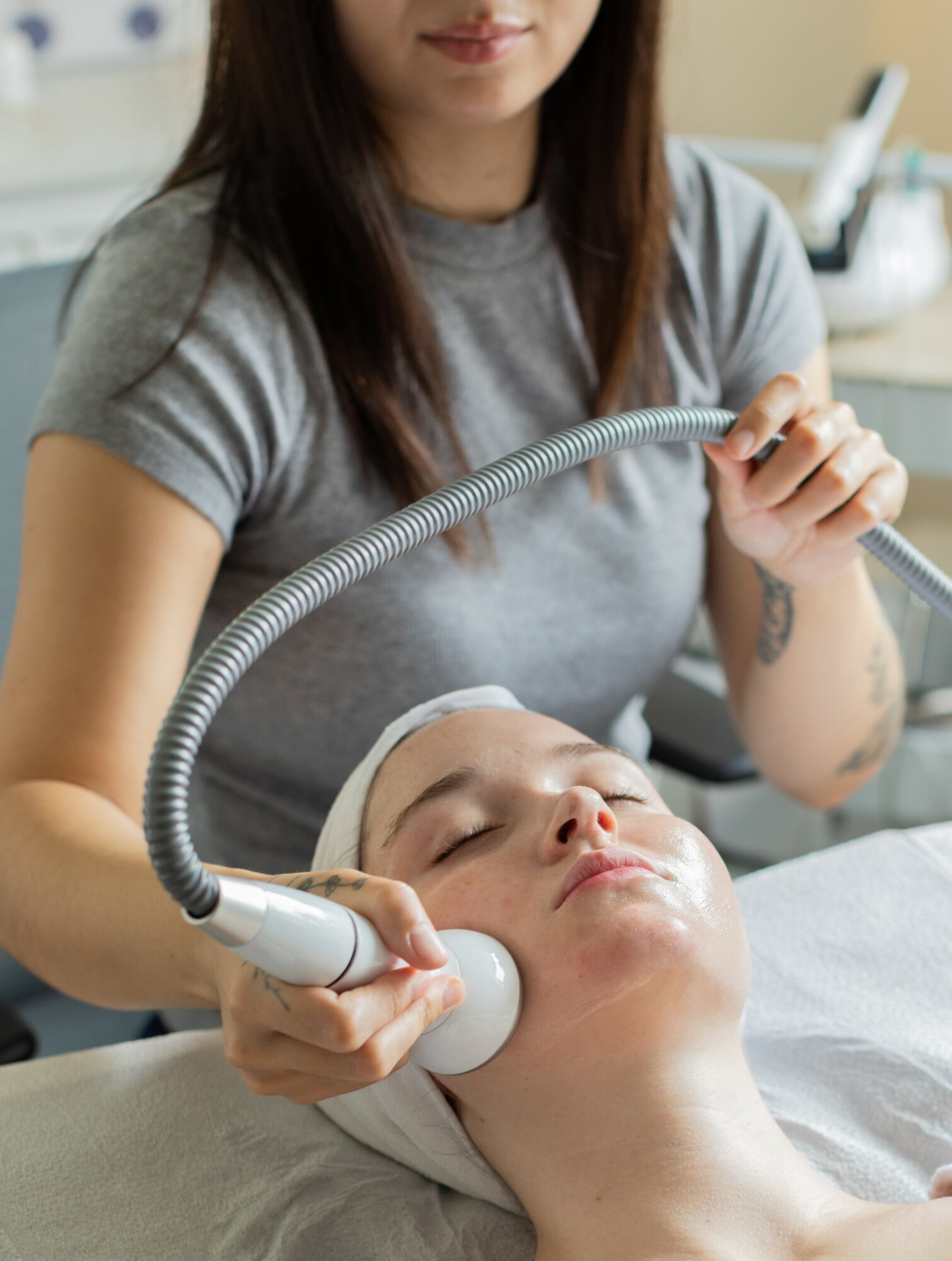 A woman receives a facial treatment with a handheld device, while a technician holds a flexible tube above her face.