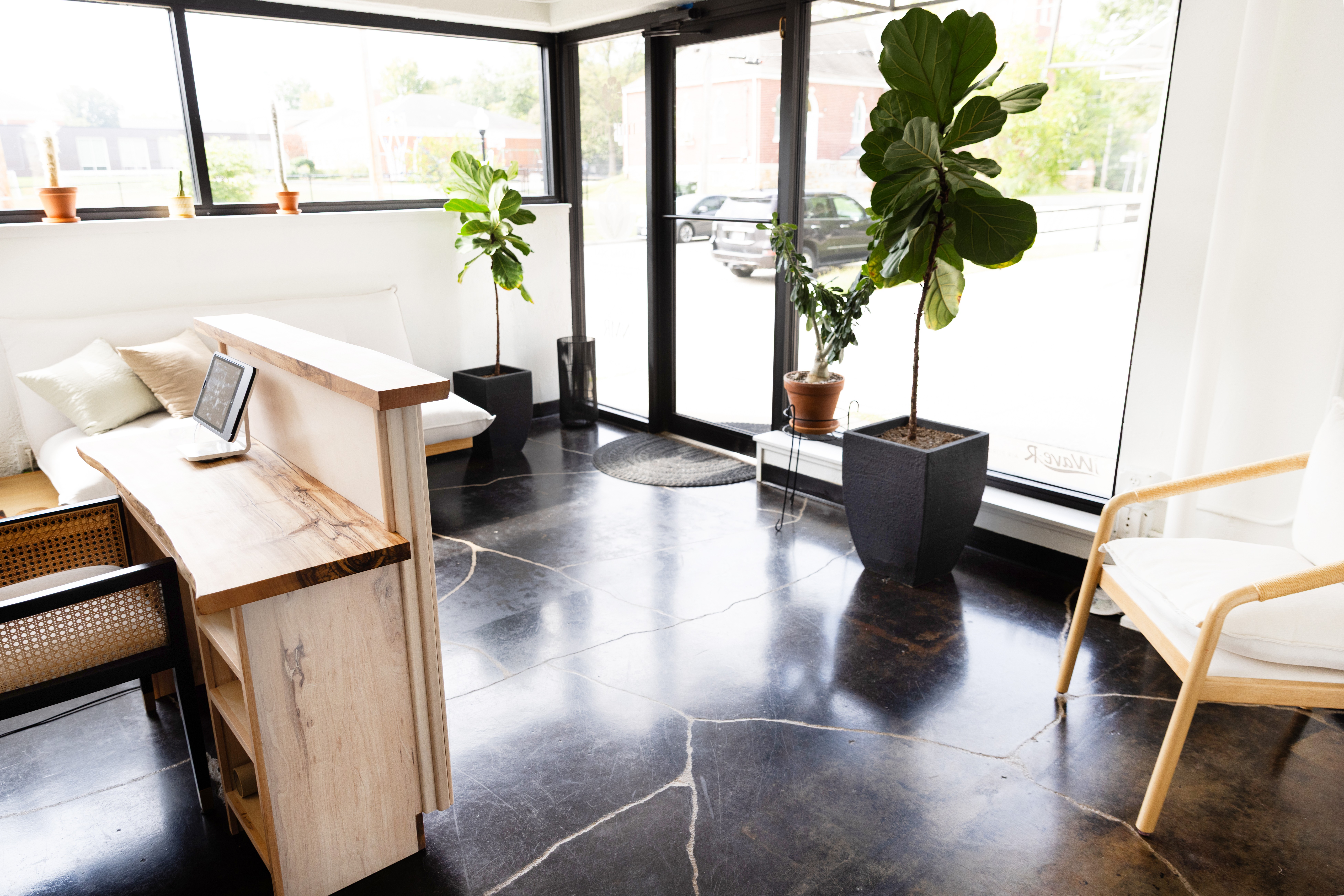 Bright room with large windows, potted plants, a wooden desk, and a white chair, with a polished dark floor.