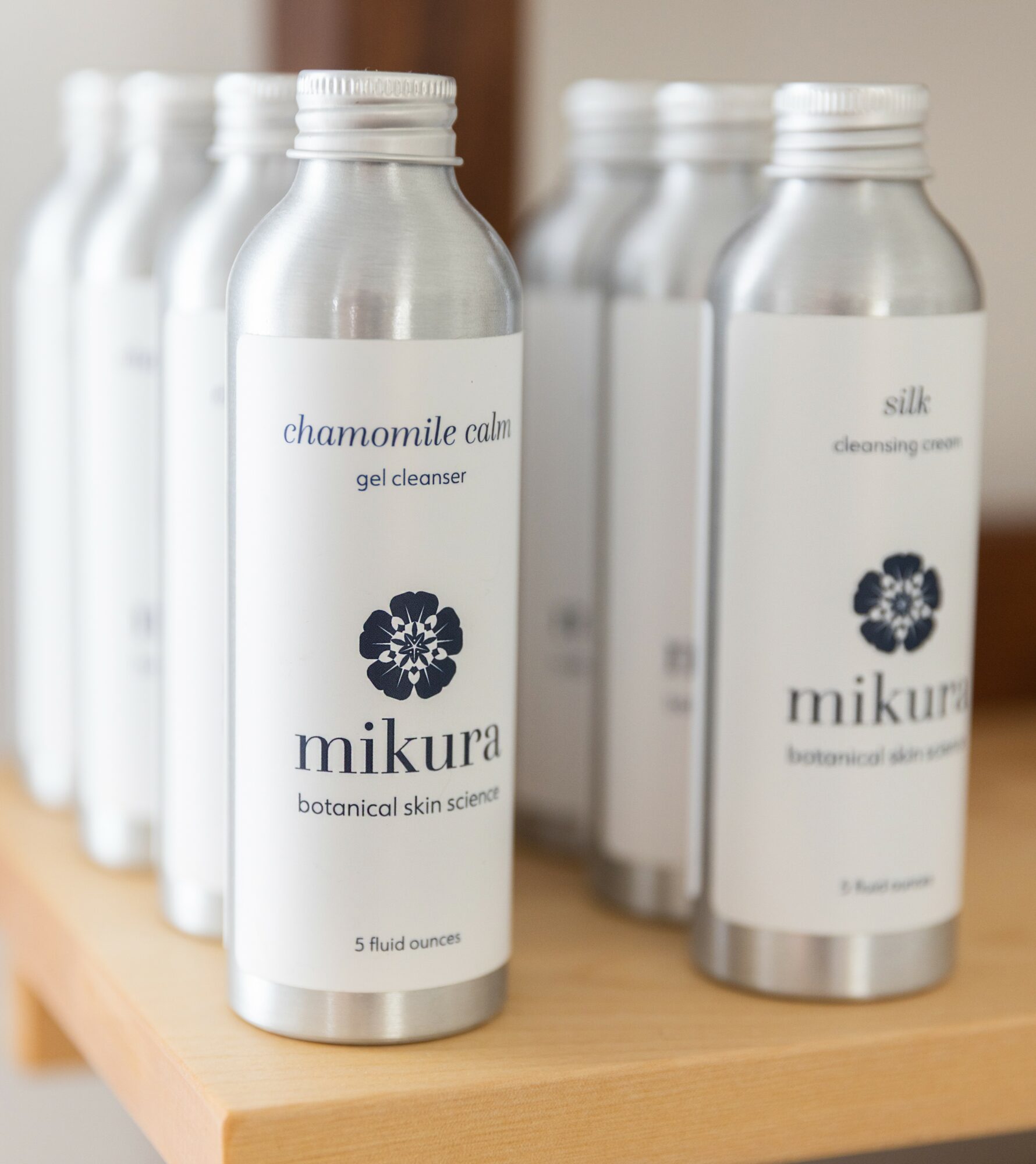 Six clear bottles with white labels and black text on a wooden surface, labeled with skincare product names, in a row.