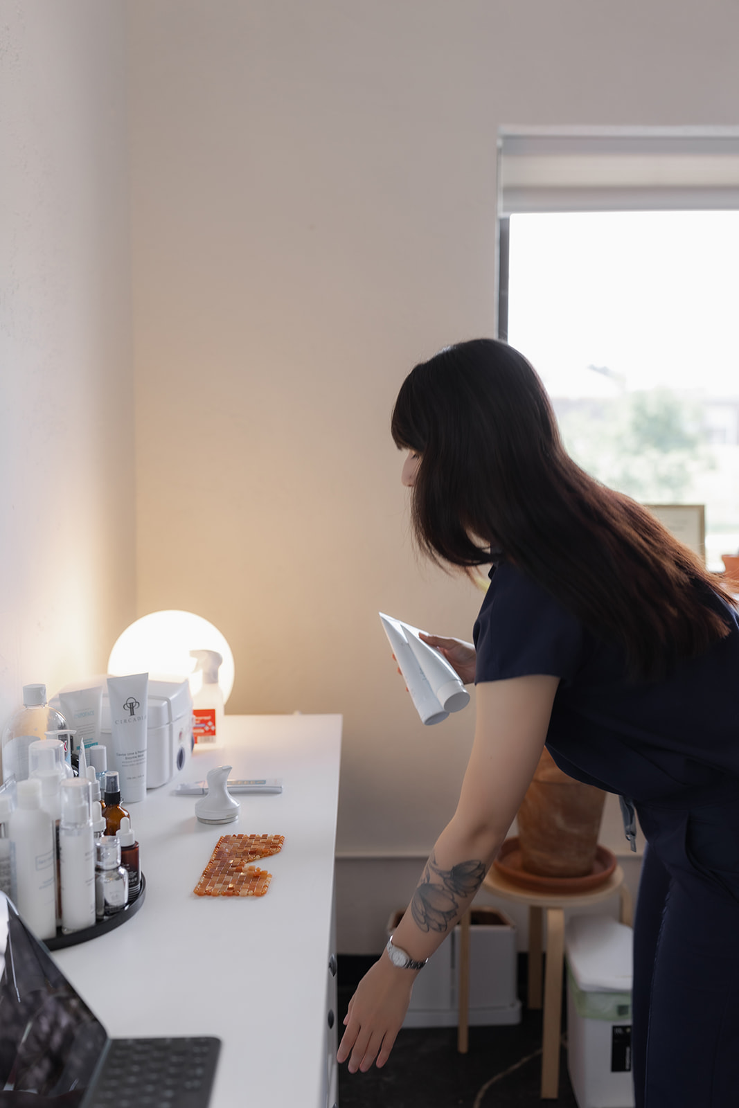 Woman with dark hair looking at a table with skincare products and a lamp, near a window.