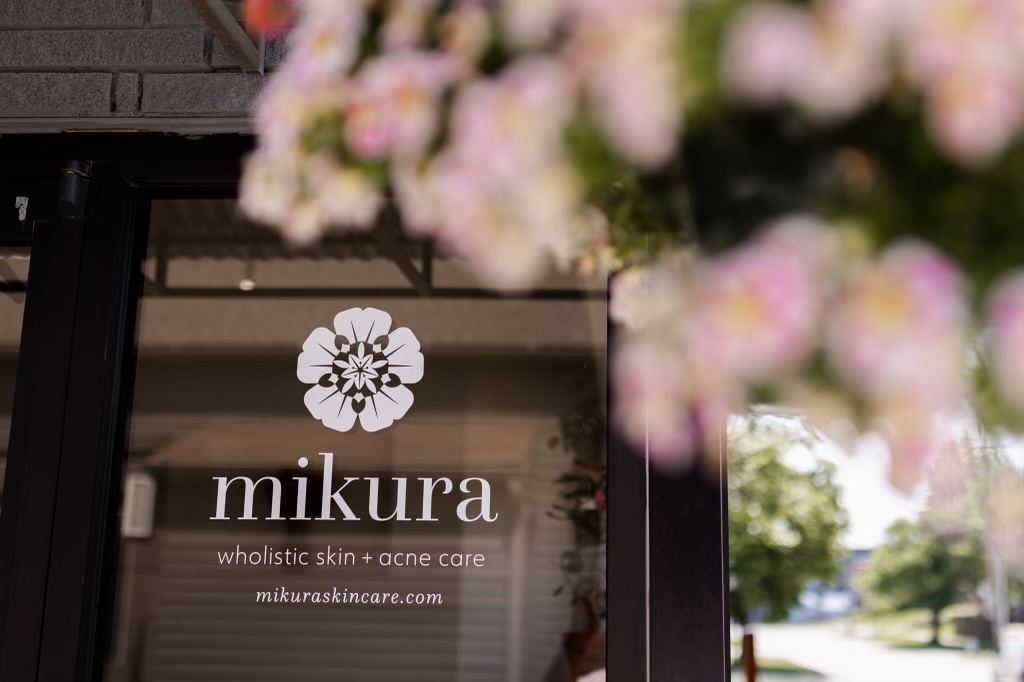 Sign for Mikura holistic skin and acne care with pink flowers in foreground, outdoor background visible.