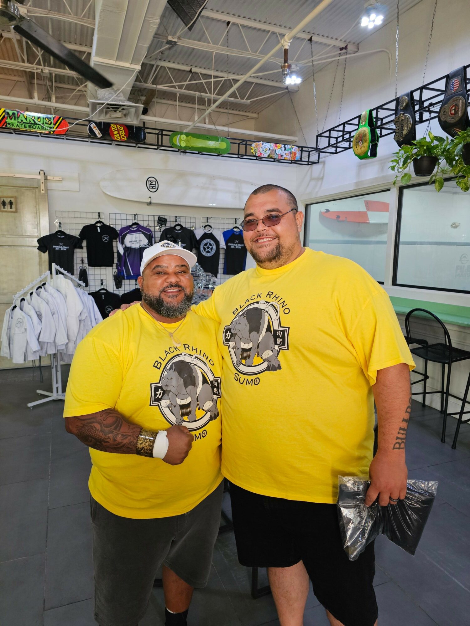 Two men in yellow shirts with a logo, one standing and one sitting, in a store with shelves and products.