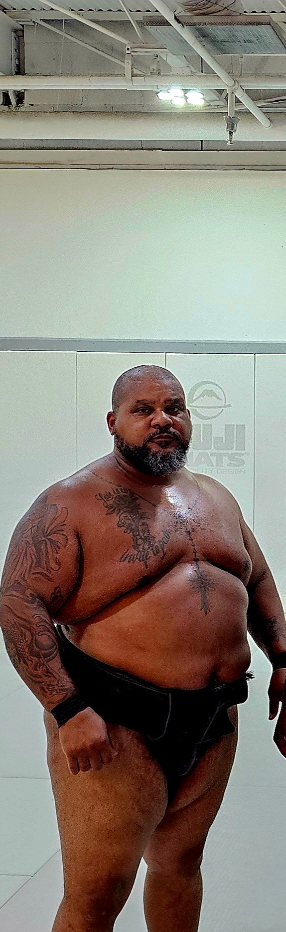 A shirtless man with tattoos standing in a room with a white wall and ceiling pipes.