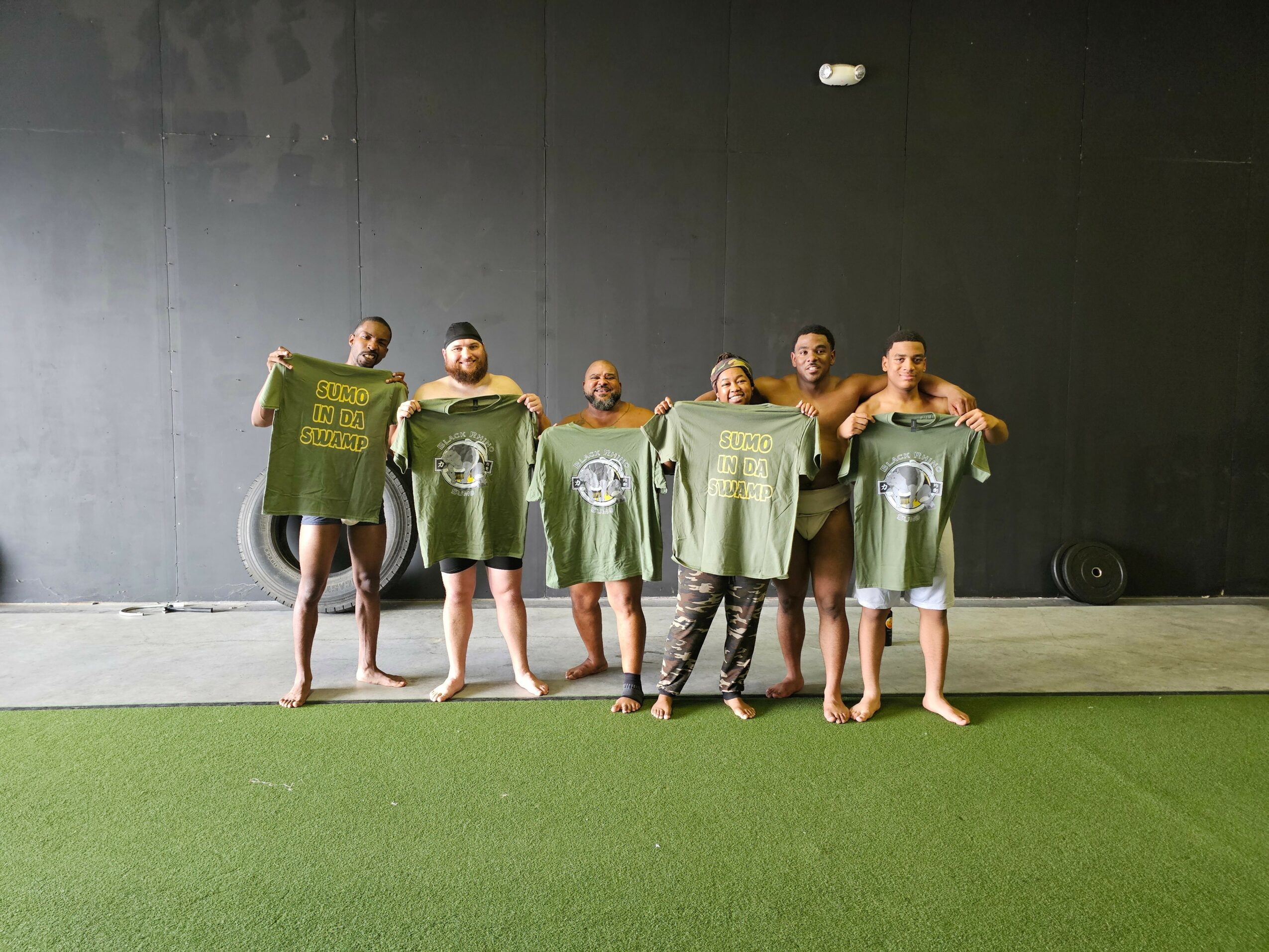 Six people standing outdoors holding green shirts, with a black wall background and green ground.