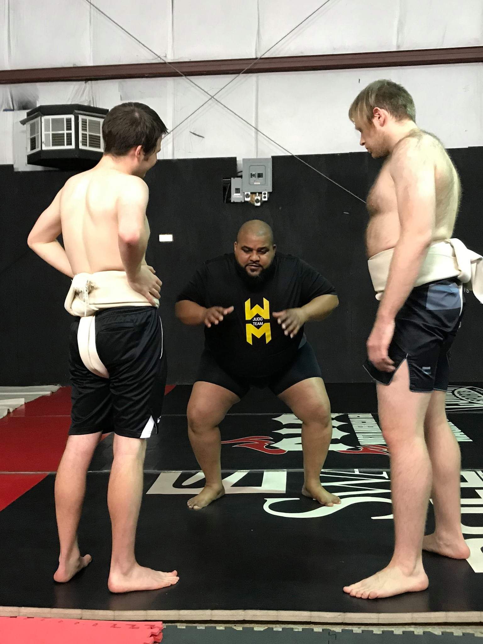 Three people in a gym, two shirtless men with shorts and a man sitting on a mat, adjusting his shorts, in front of a black wall.