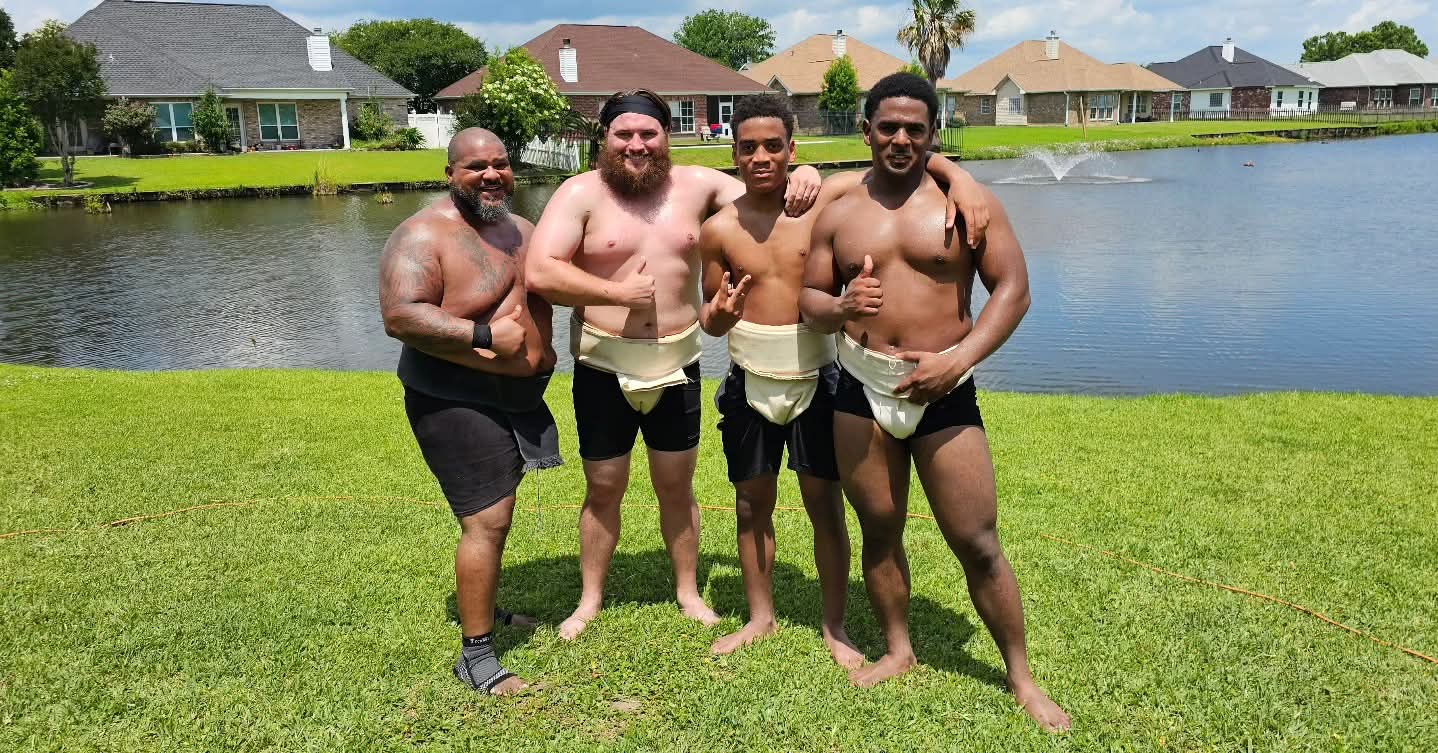 Four men stand on grass near water, smiling and posing for a photo, with houses in the background.