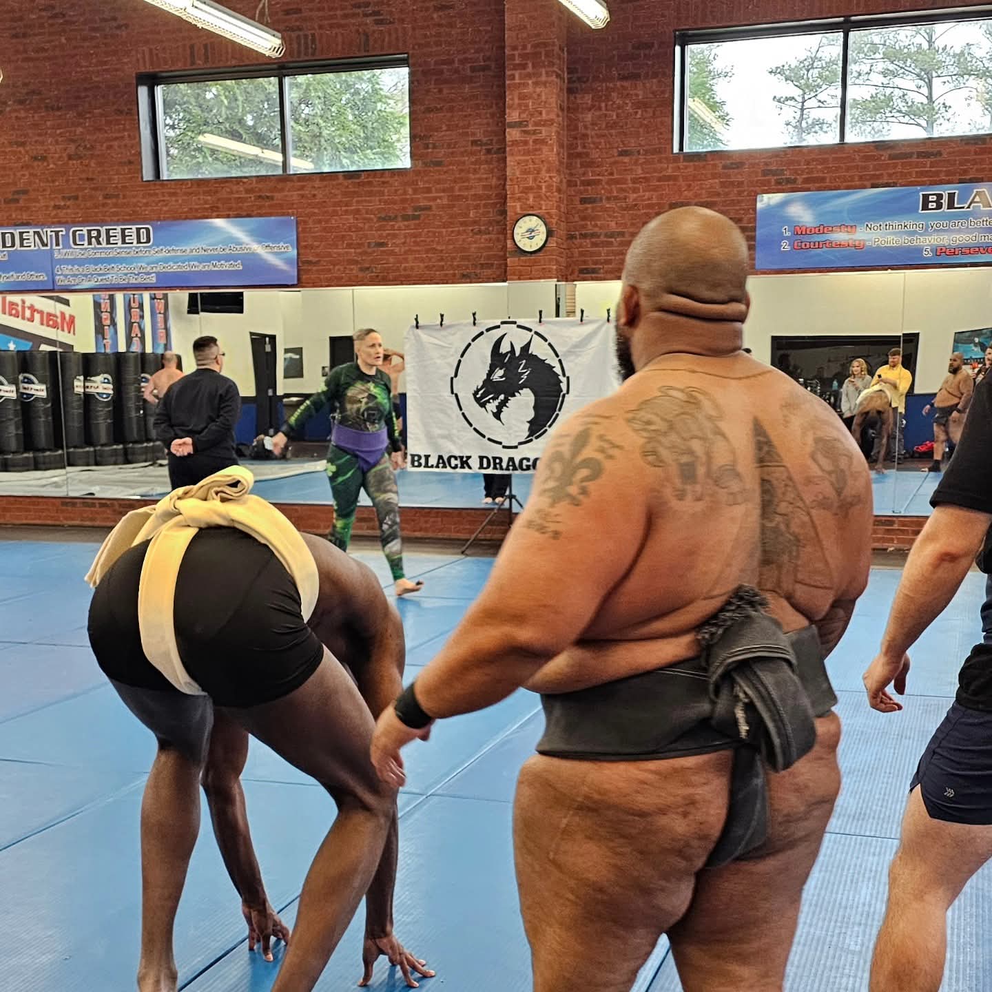 Two men in a martial arts gym, one bending over and the other with a tattooed back, surrounded by people and training equipment.