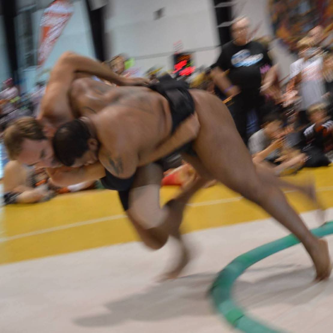 Two wrestlers engaged in a match on a yellow mat, with spectators watching in the background.