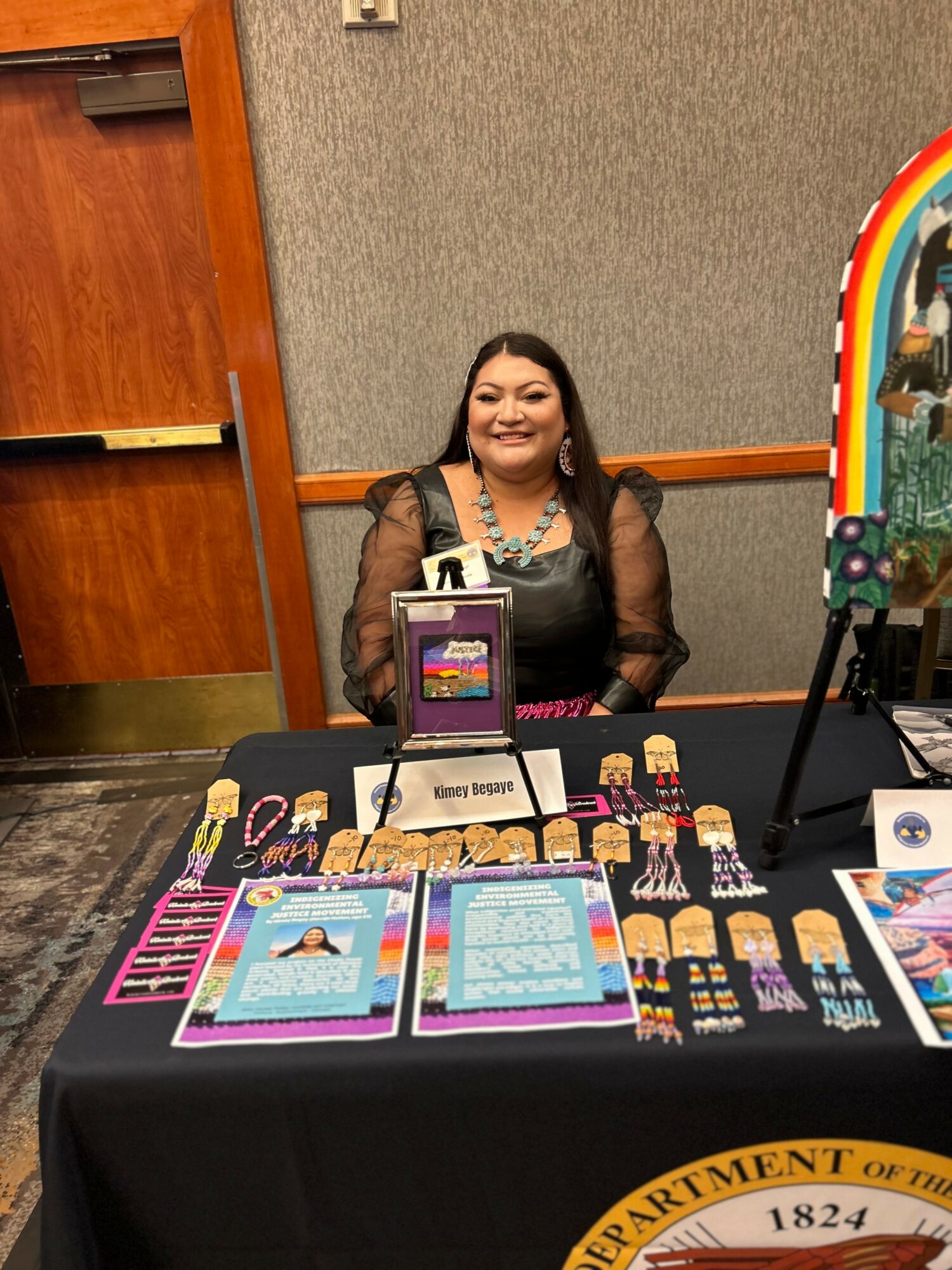Smiling woman sitting behind a table with colorful brochures and jewelry displays, in a room with gray walls and a rainbow decoration.