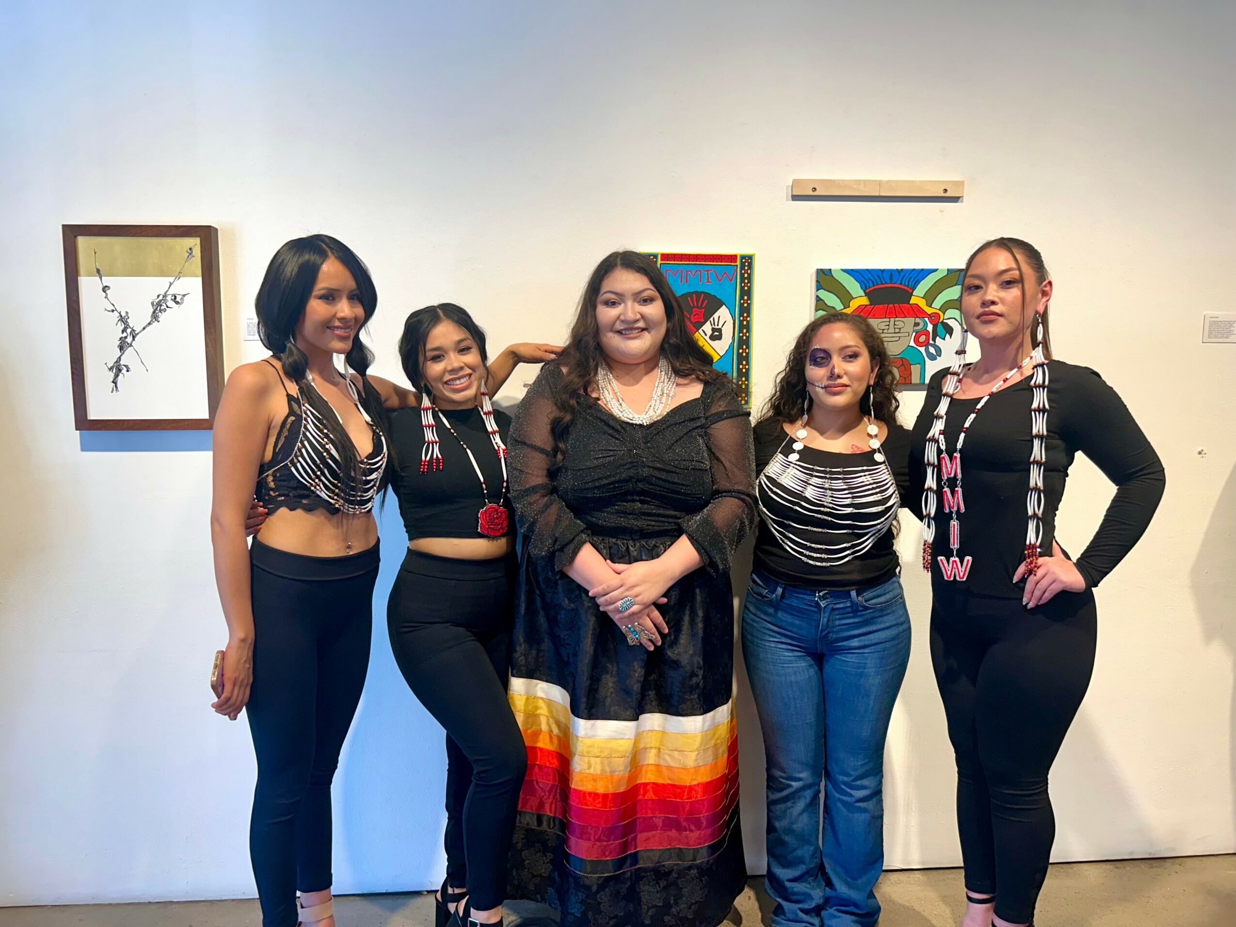 Five women standing together in an art gallery, smiling, with colorful artwork on the wall behind them.