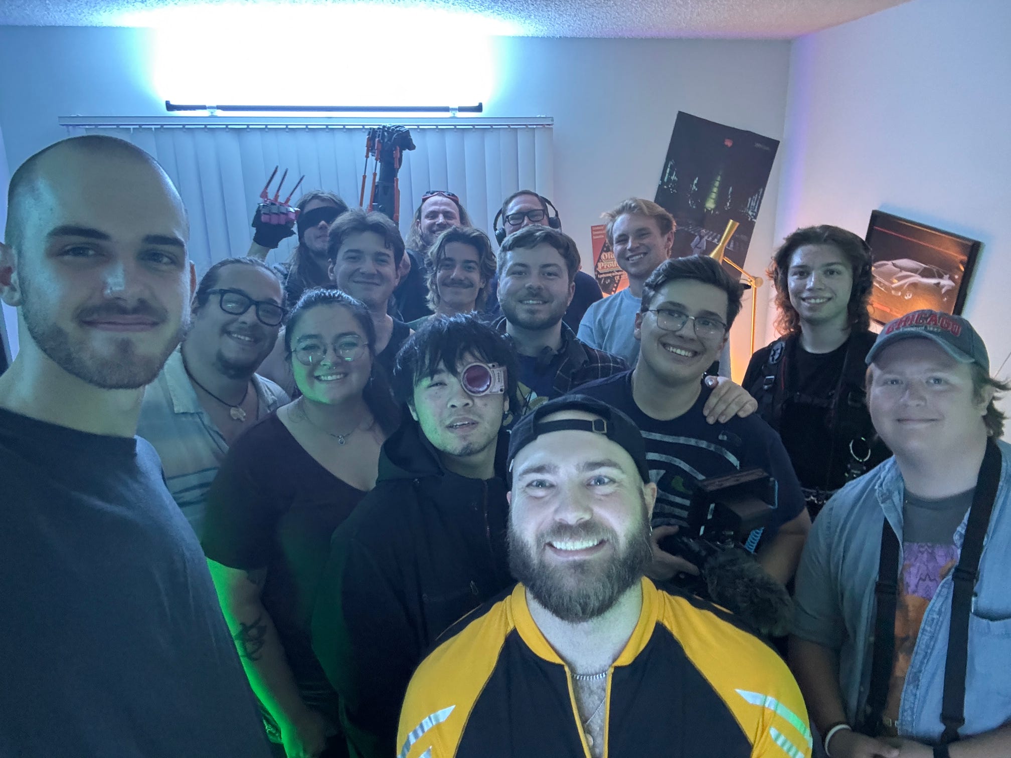Group of people smiling indoors, some wearing glasses, with a bright light and posters in the background.