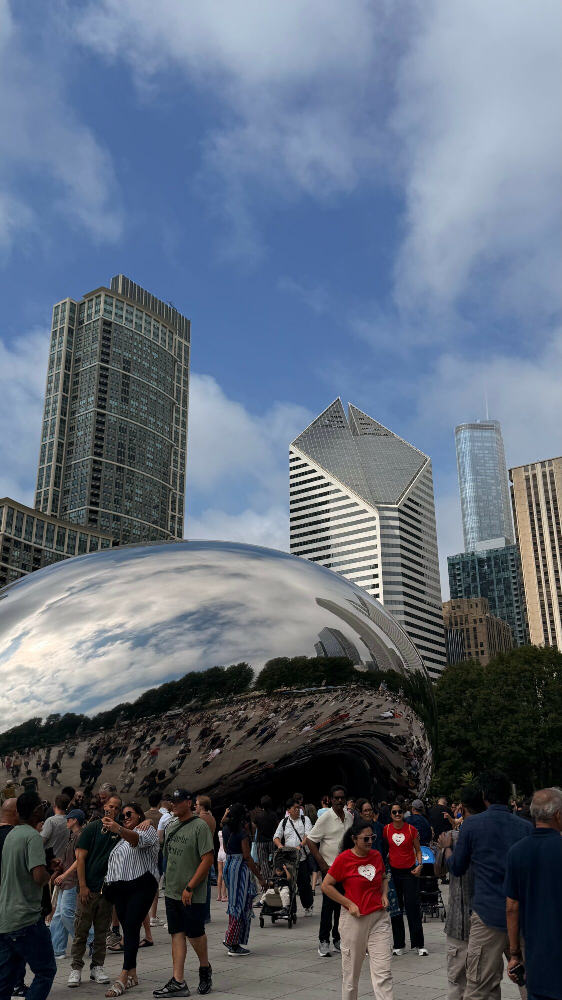People gather in a city park with tall buildings and a reflective sculpture under a partly cloudy sky.