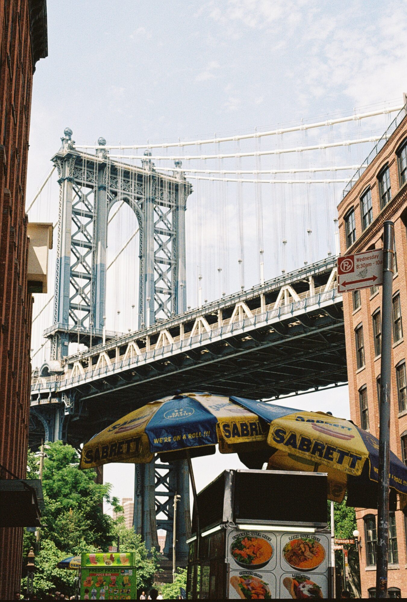 View of a bridge with a street vendor stand and umbrellas in foreground, buildings on sides, and sky above.
