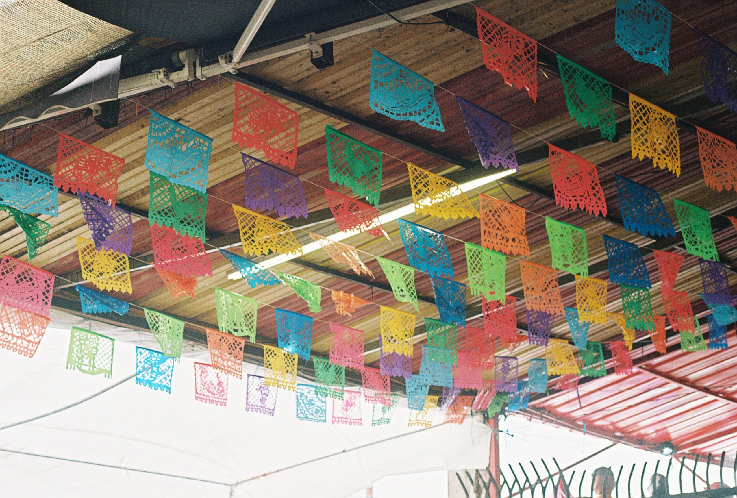 Colorful papel picado banners hanging under a roof, with a fence visible in the background.