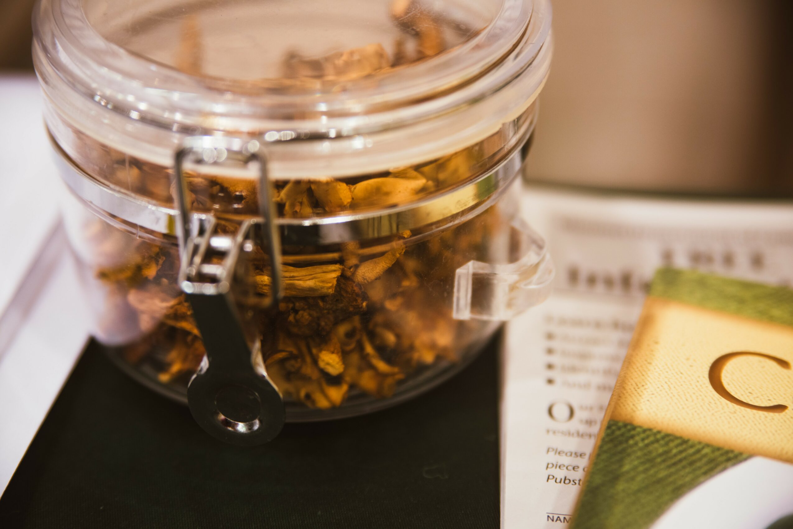 Clear jar filled with dried flower petals or herbs, placed on a dark surface with a paper and a book nearby.