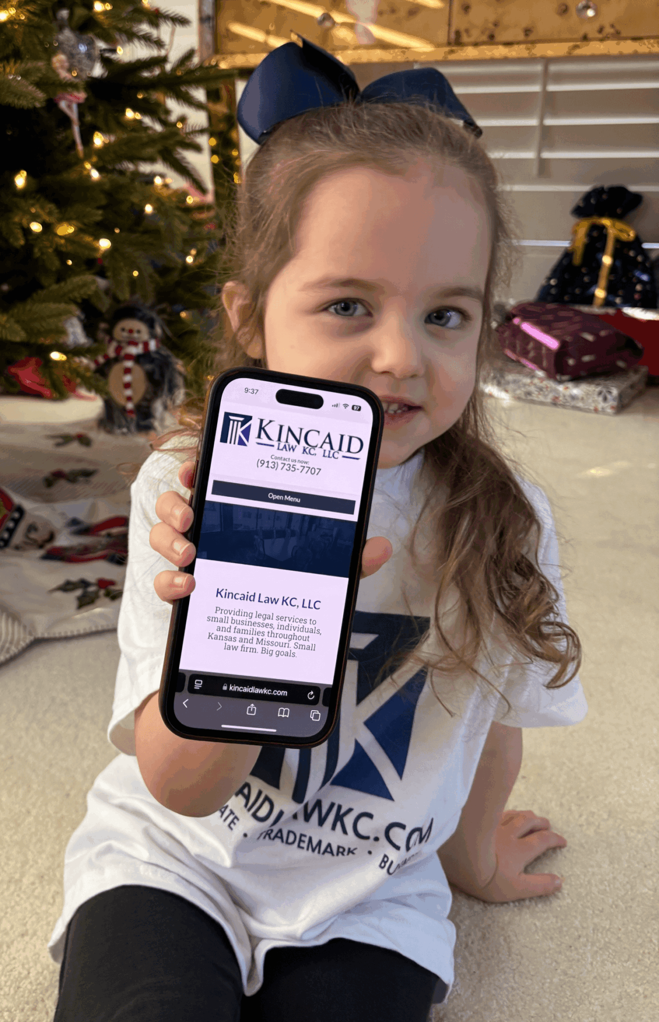 Young girl with long hair and a blue bow holding a phone displaying a business card, sitting on a carpeted floor with Christmas decorations.
