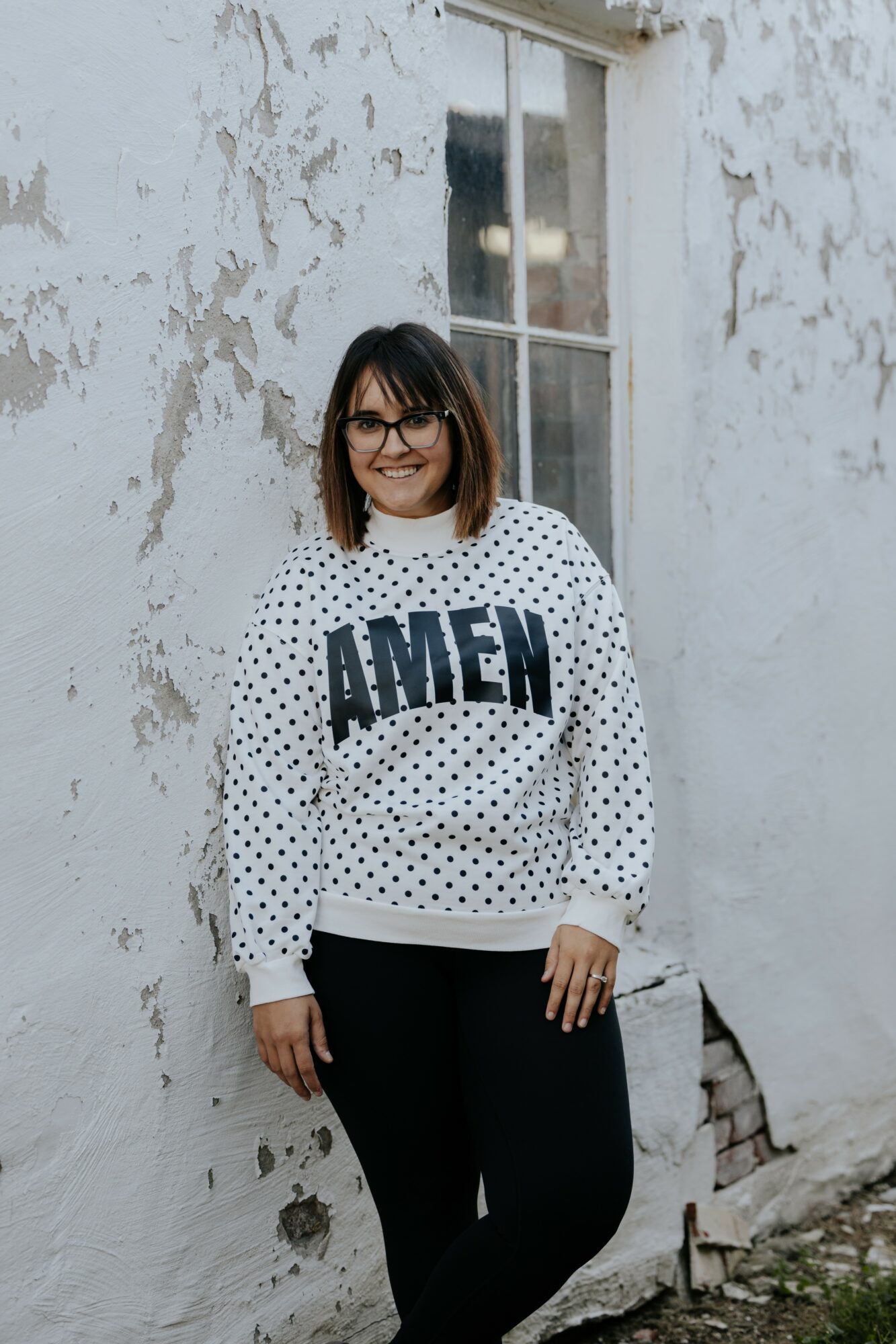 Woman with glasses and shoulder-length hair standing outdoors against a weathered white wall, smiling, wearing a white sweater with black polka dots and 'AMEN' text.