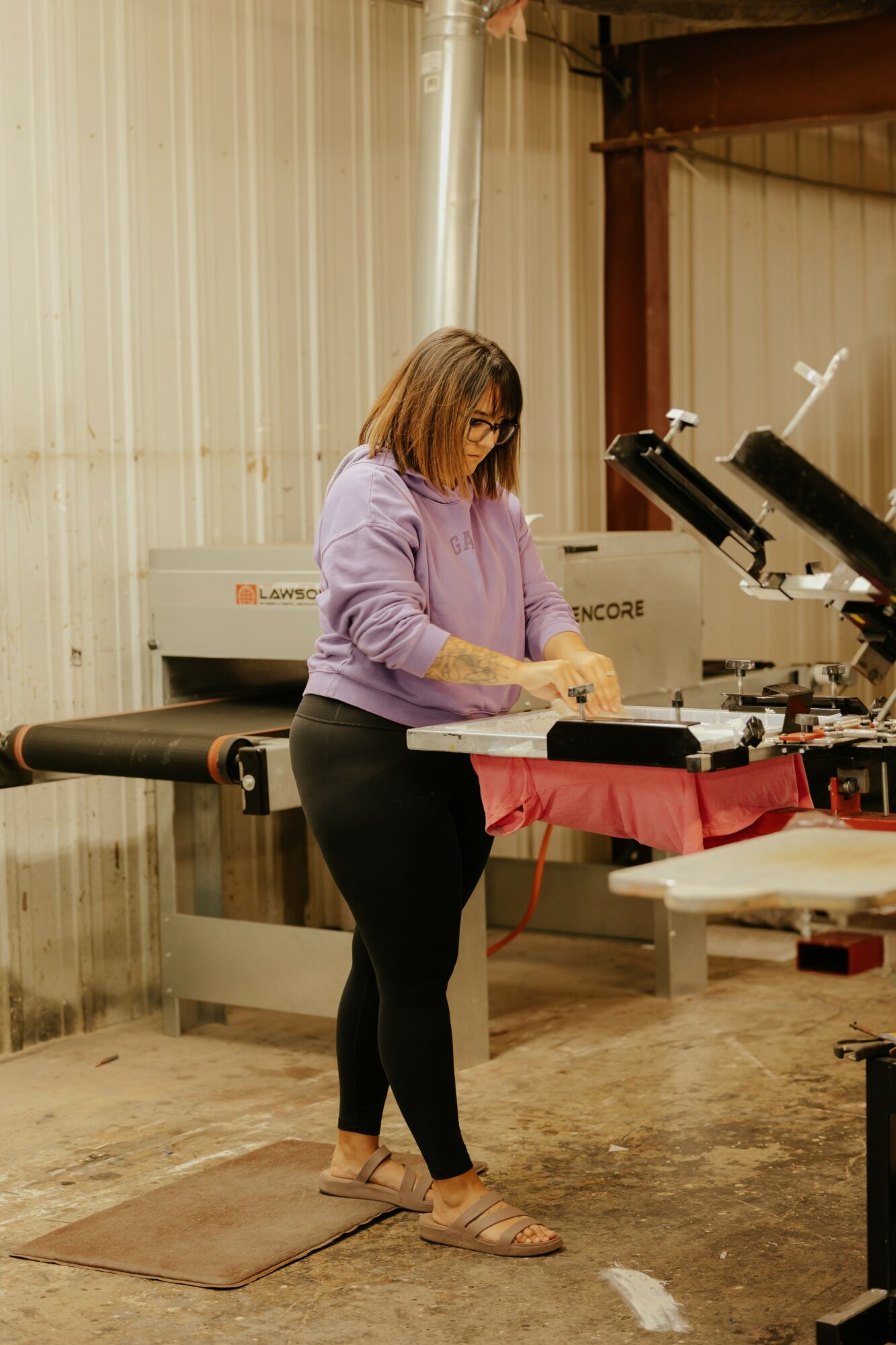Woman in purple sweater working with equipment in a workshop, standing on a mat, with machinery and tools around.