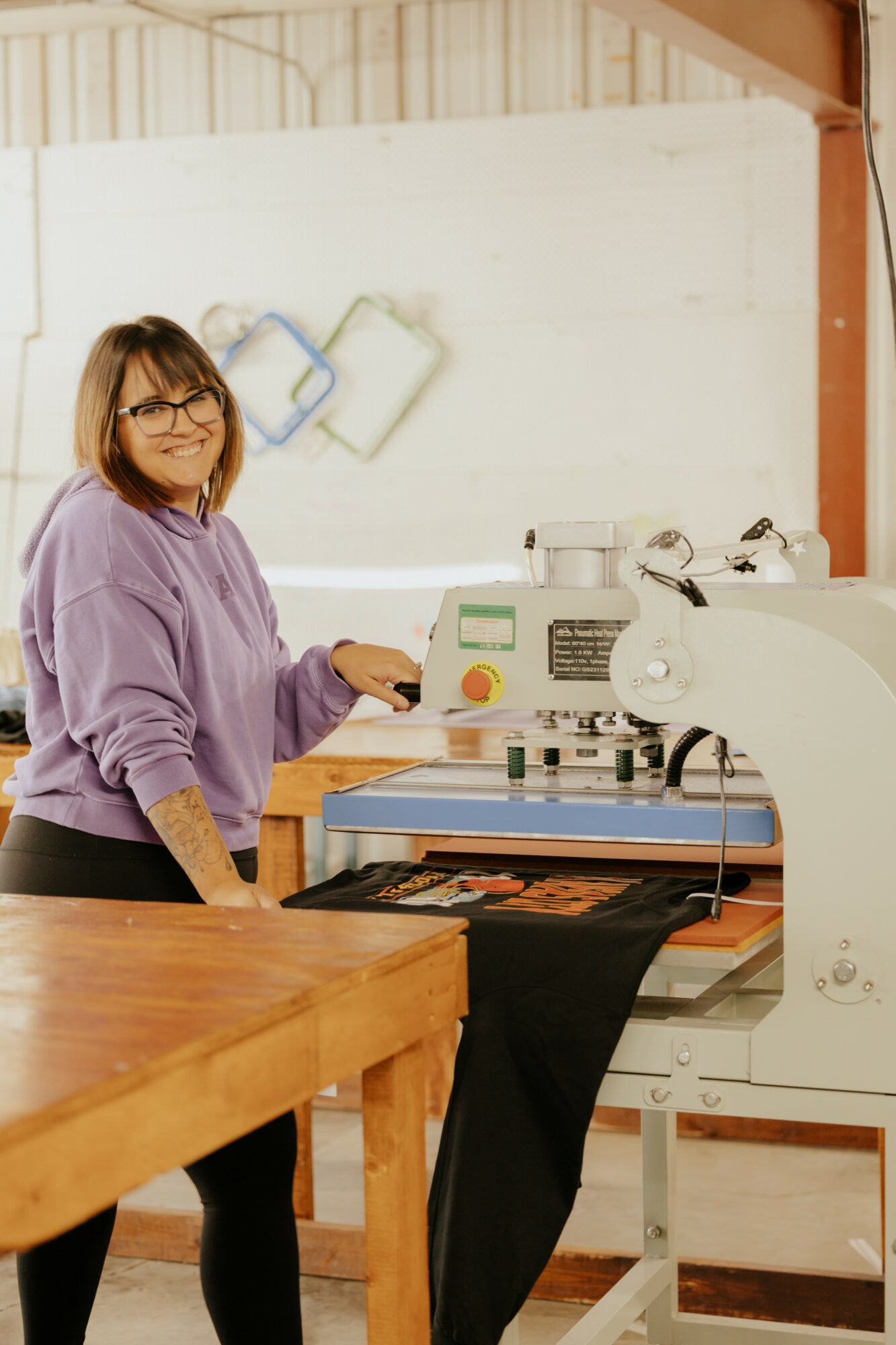 Woman in purple sweater working with a machine in a workshop, smiling, with a white wall and equipment in background.