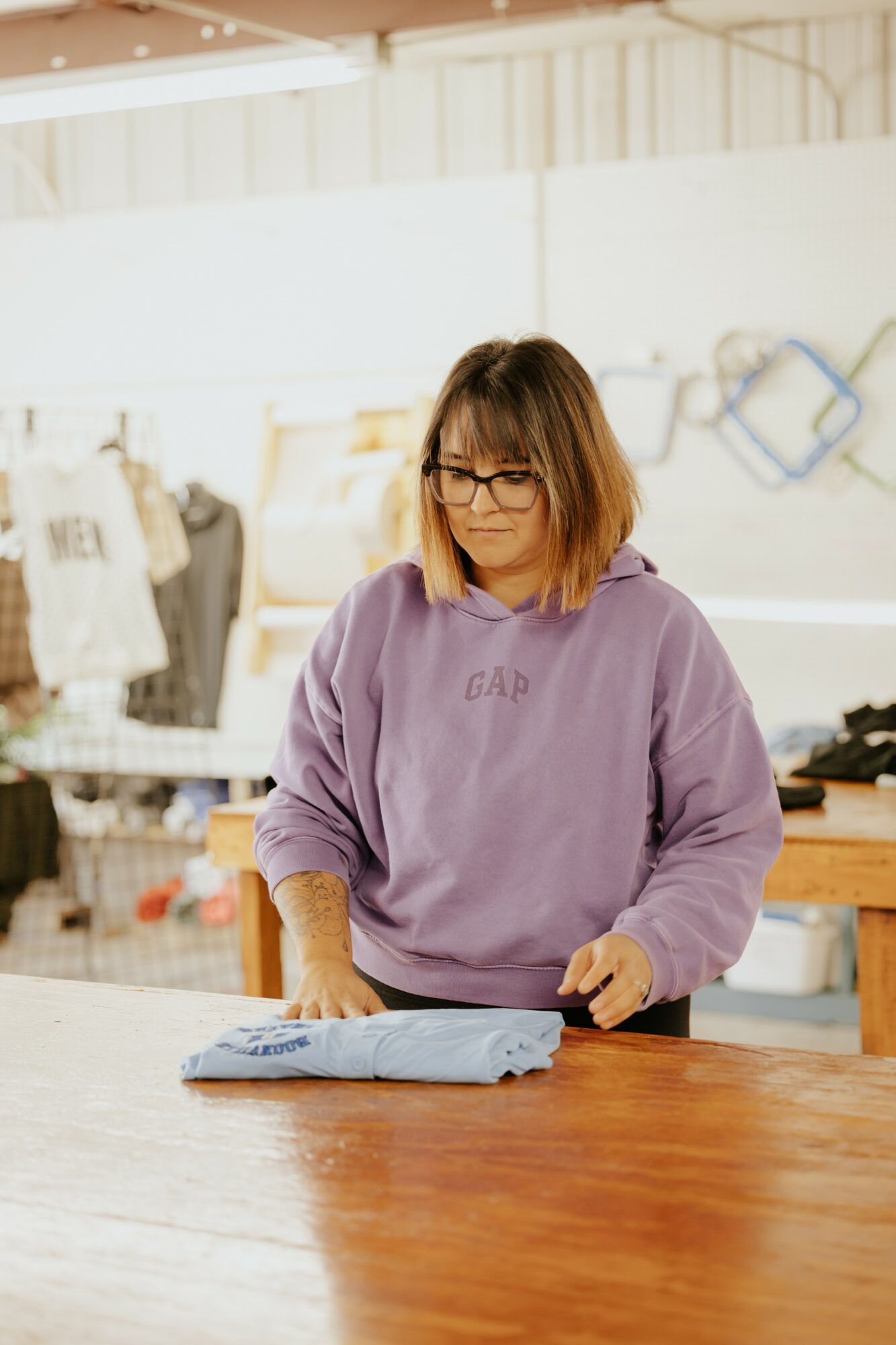 Woman with glasses folding clothes at a table in a store, wearing a purple hoodie, with clothing displays in background.