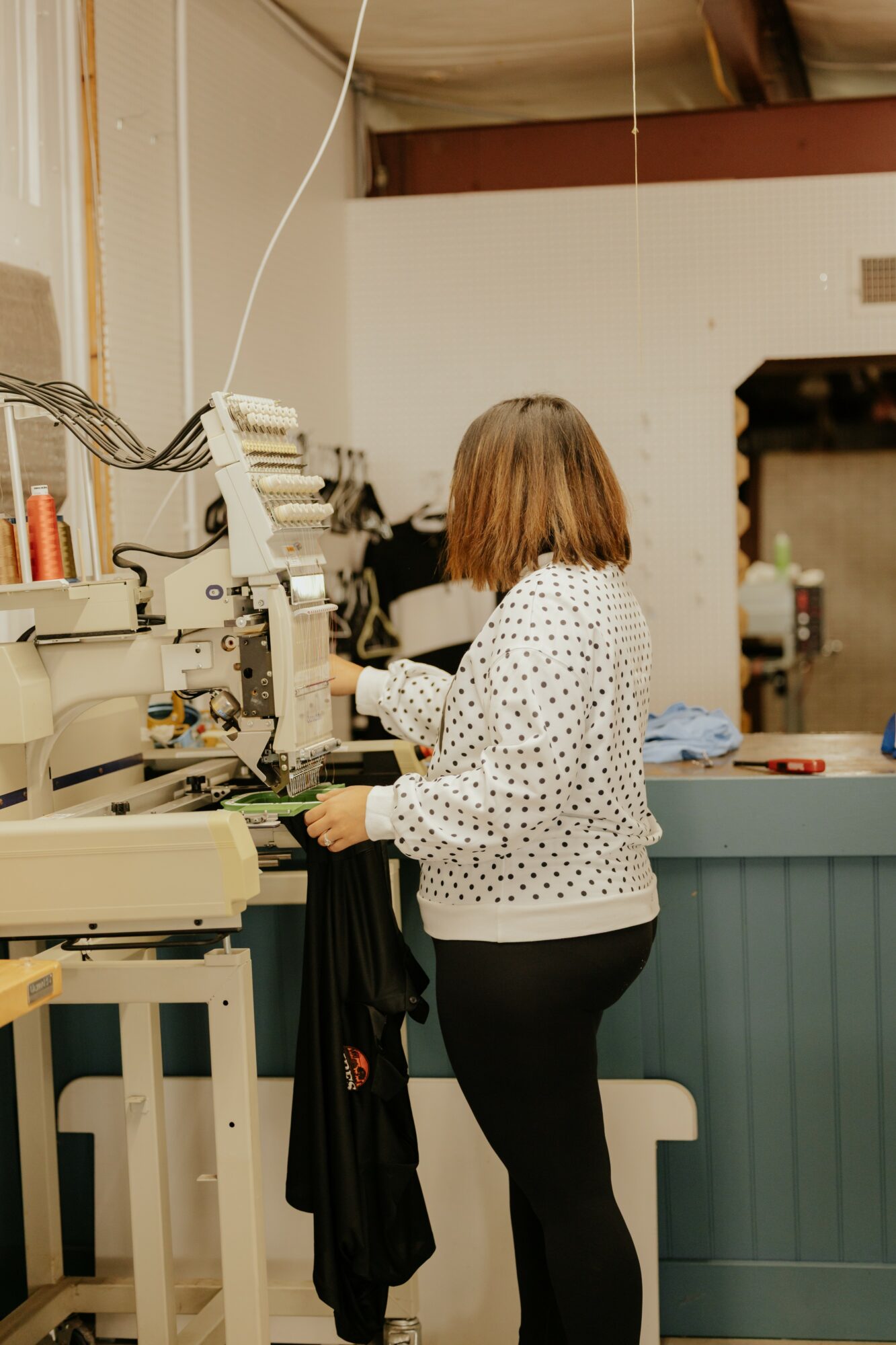 Woman with shoulder-length hair working at a machine in a workshop, wearing a white polka-dot blouse and black pants.