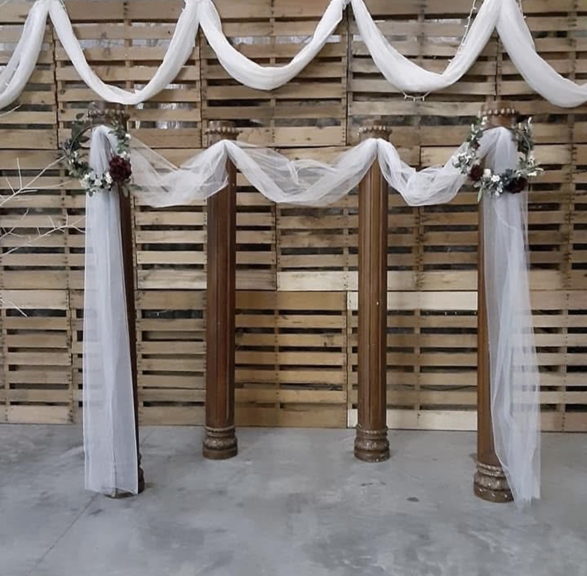 Decorative wedding arch with white fabric and floral arrangements on wooden posts, against a wooden backdrop.