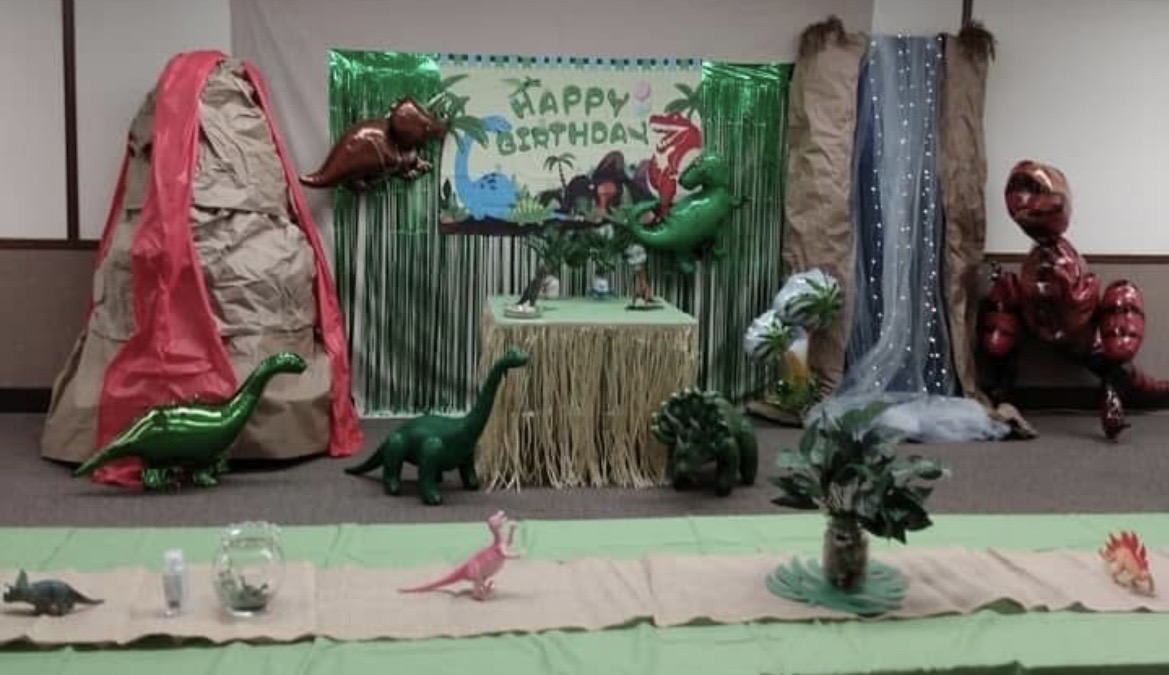 Decorative birthday scene with dinosaur-themed balloons, a green backdrop, and a table with a 'Happy Birthday' sign.