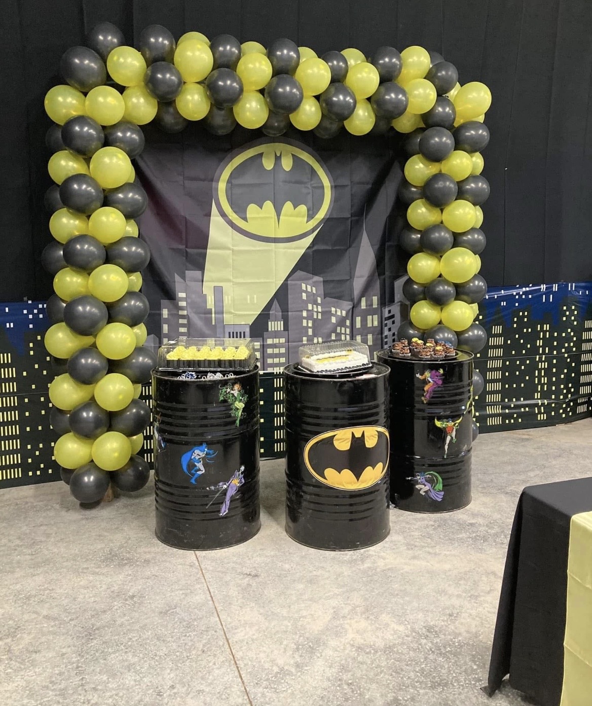 Decorative Batman-themed setup with black and yellow balloons, black barrels with Batman logos, and cityscape backdrop.