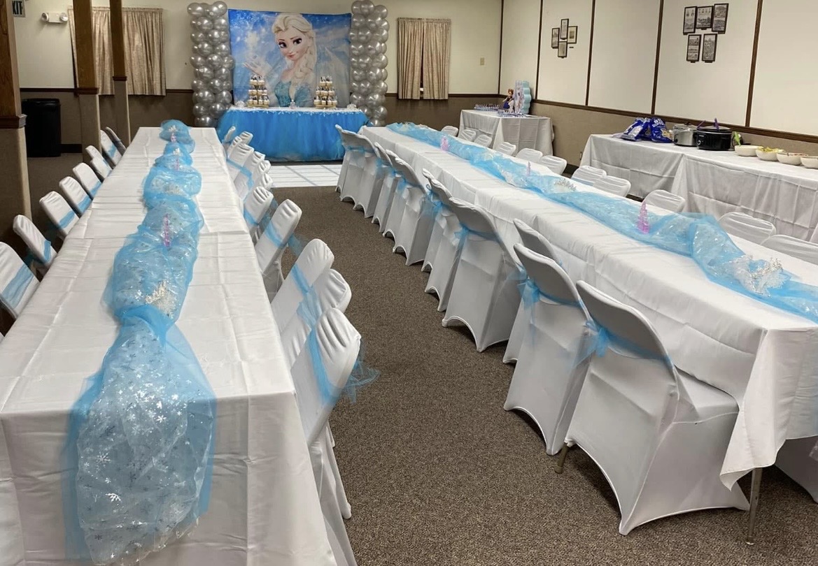 Long banquet tables with white tablecloths and chairs, decorated with blue and clear plastic coverings, in a decorated event room.