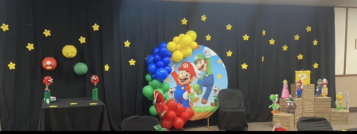 Decorated stage with balloons, stars, and a large Mario-themed backdrop, likely for a children's party or event.