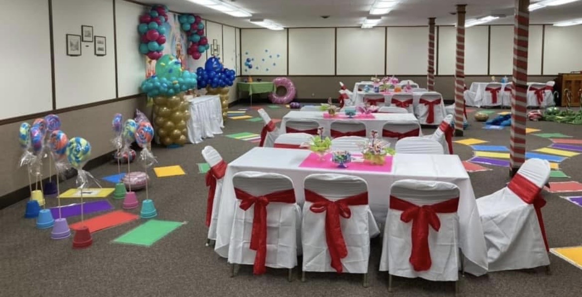 Party room decorated with balloons, tables with pink and white decorations, and chairs with red bows, ready for celebration.