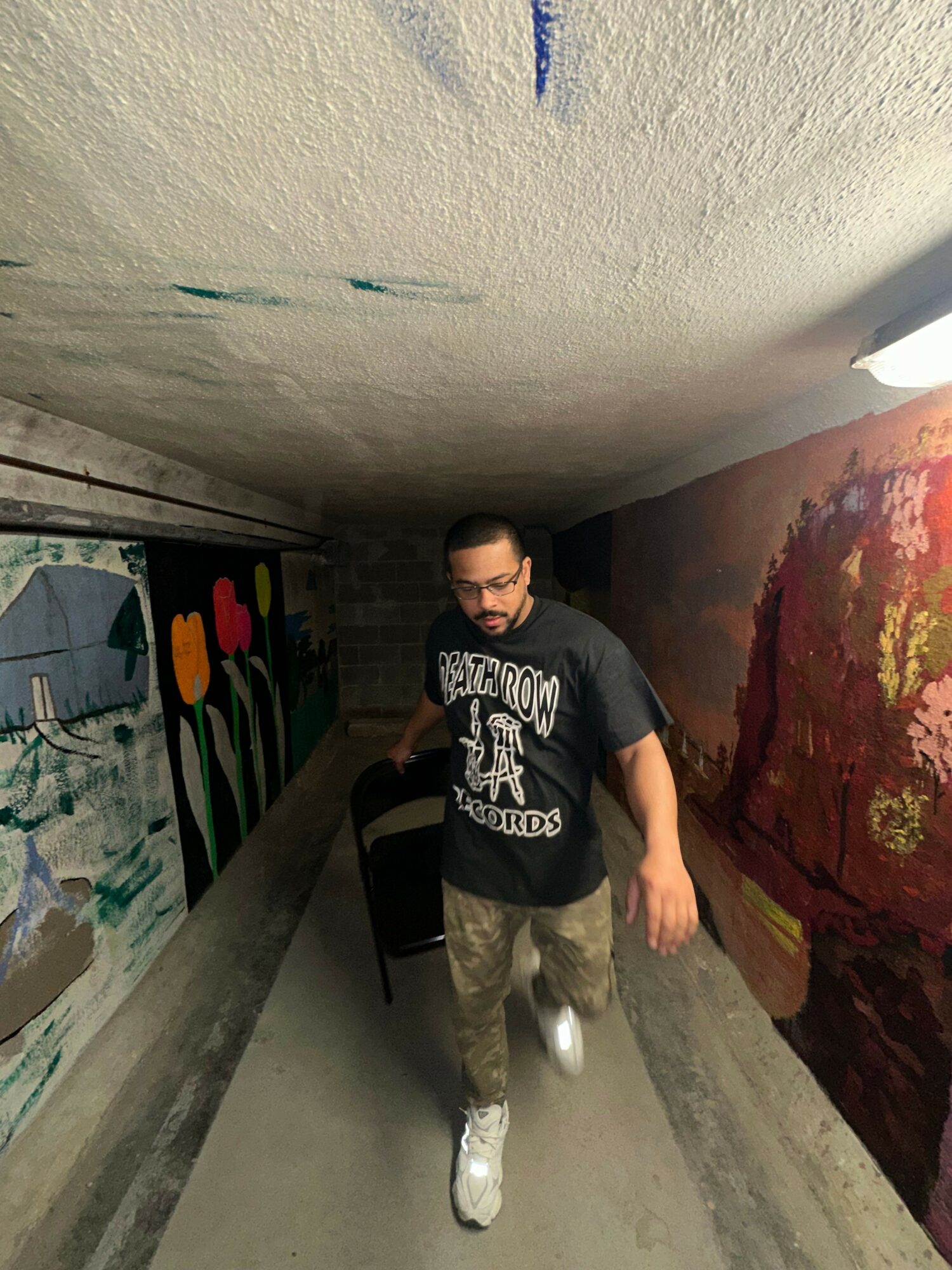 Person walking in a narrow corridor with colorful artwork on the walls, wearing glasses, a black hoodie, and camouflage pants.