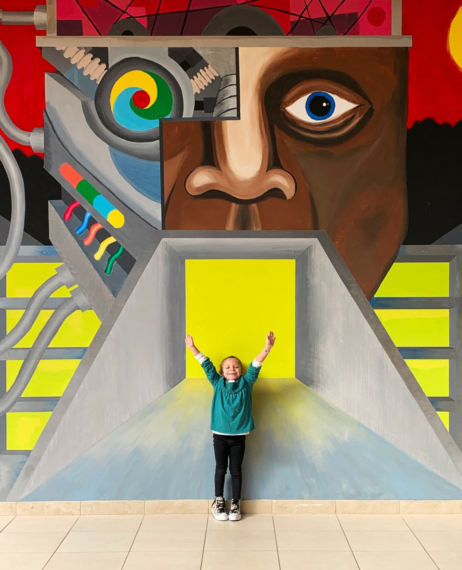 Child standing with arms raised in front of a colorful mural with a large face and abstract shapes.