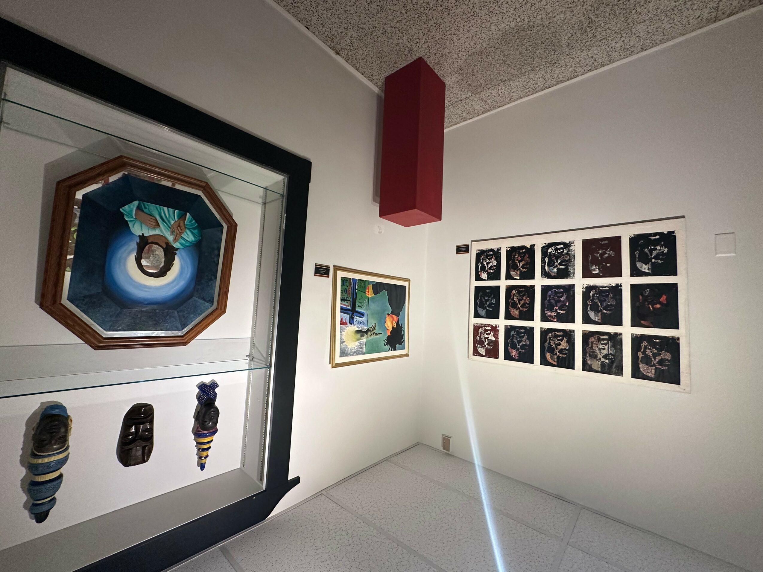 Gallery corner with framed artwork on white walls, including a grid of small images and a colorful piece, with a red column.