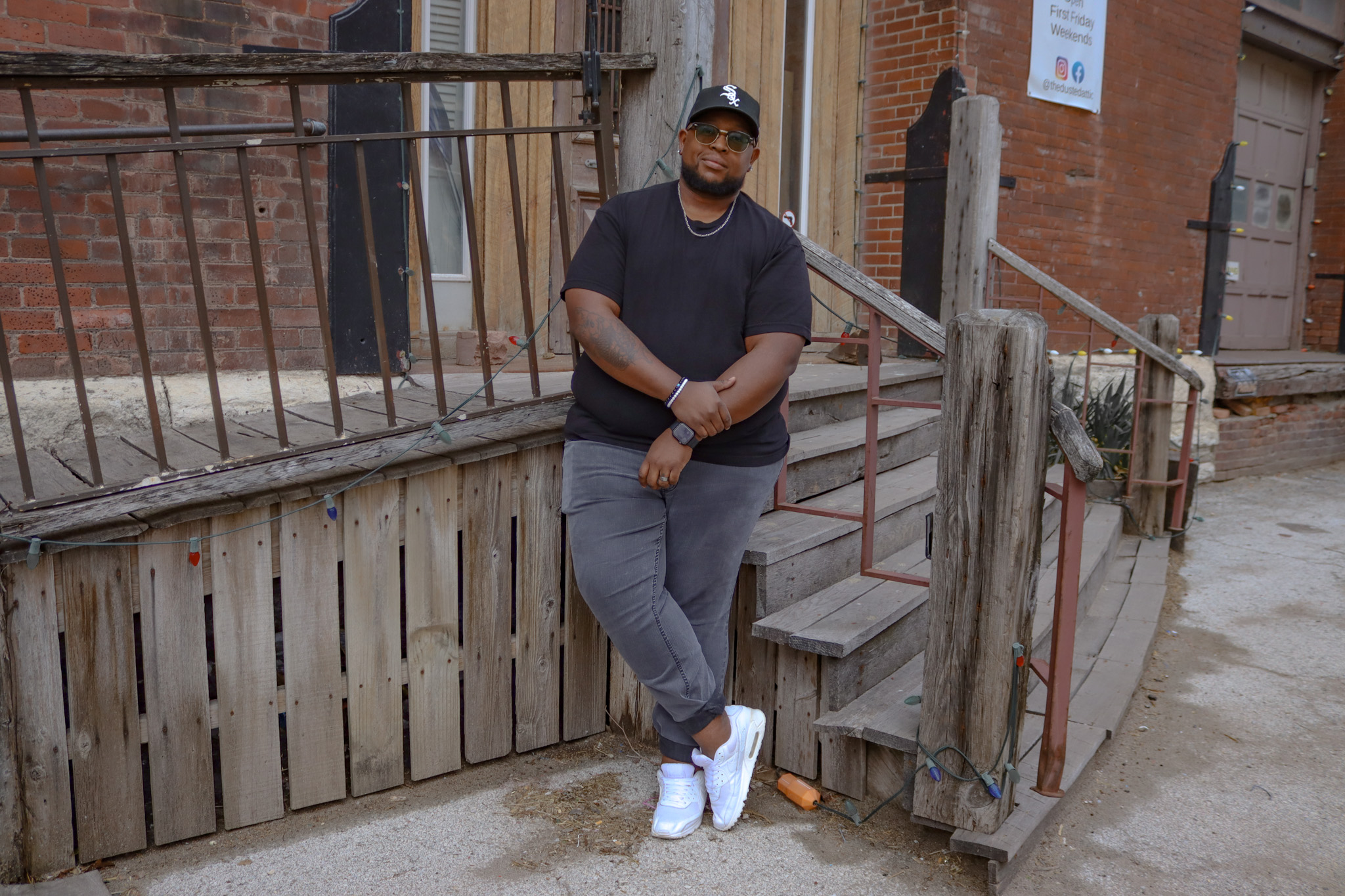 Person standing outdoors near a wooden fence and stairs, wearing a black shirt, gray pants, white shoes, and a cap.
