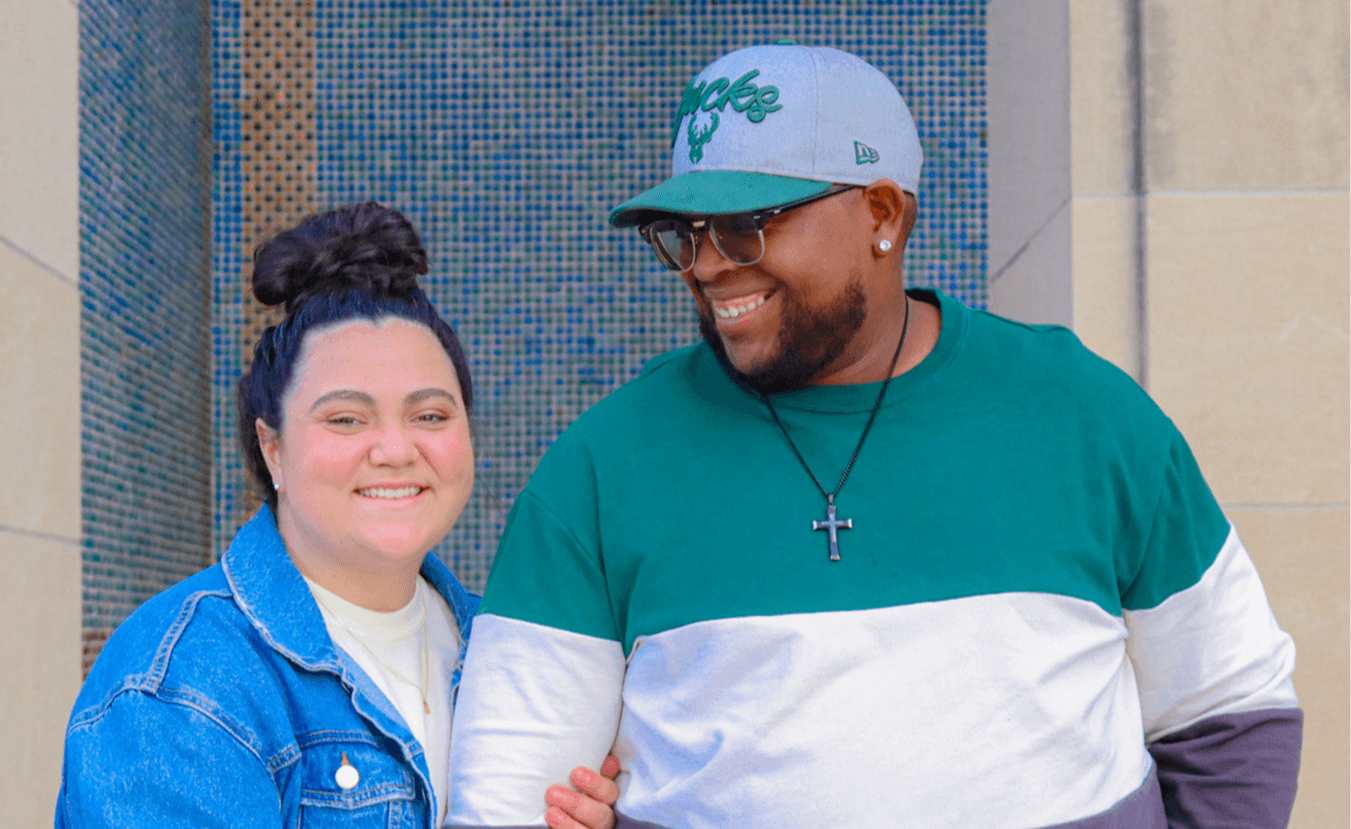 Two people smiling, one wearing a denim jacket and the other in a green and white shirt with a cap, standing outdoors.