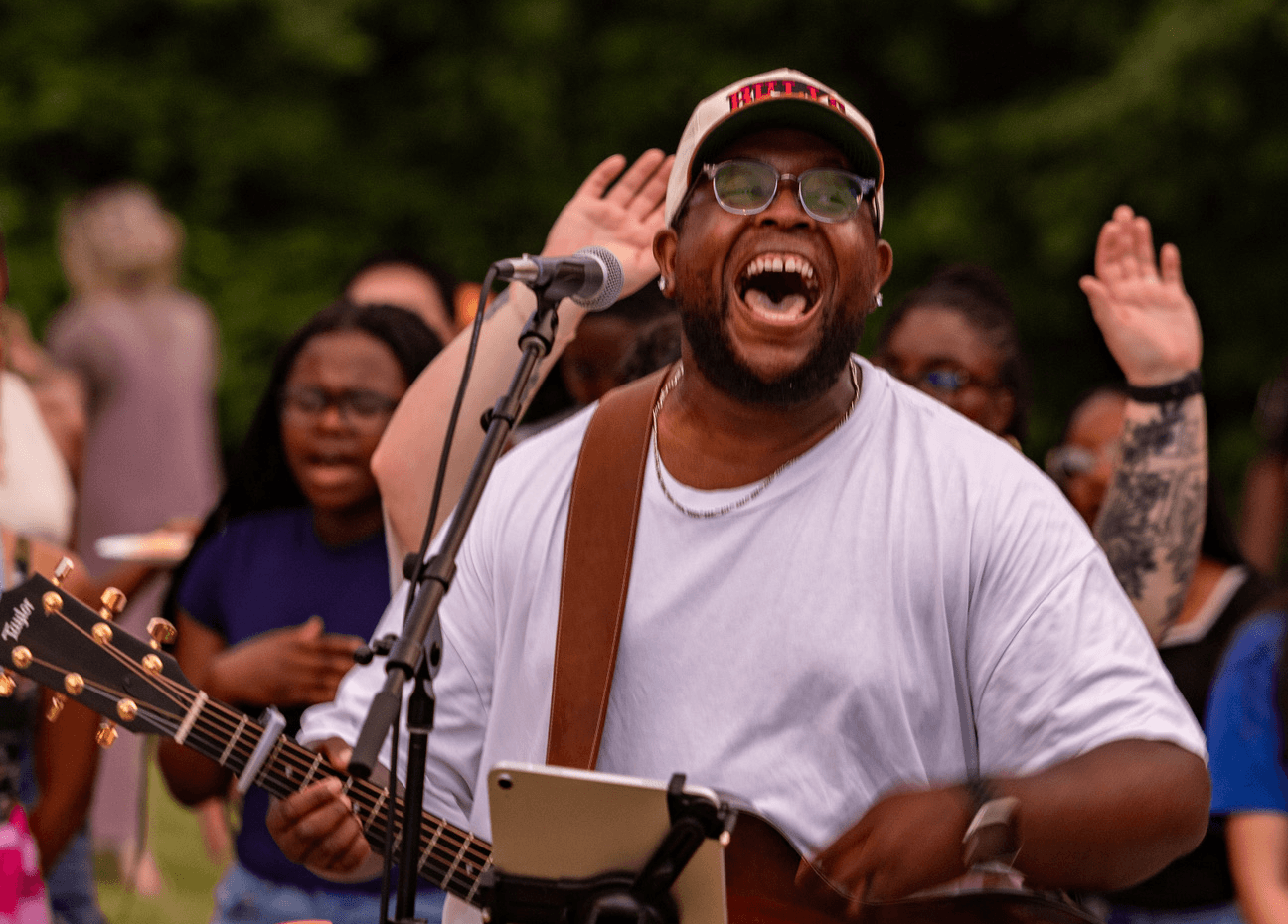 Man singing and playing guitar outdoors with a group of people, some raising hands, in a park setting.