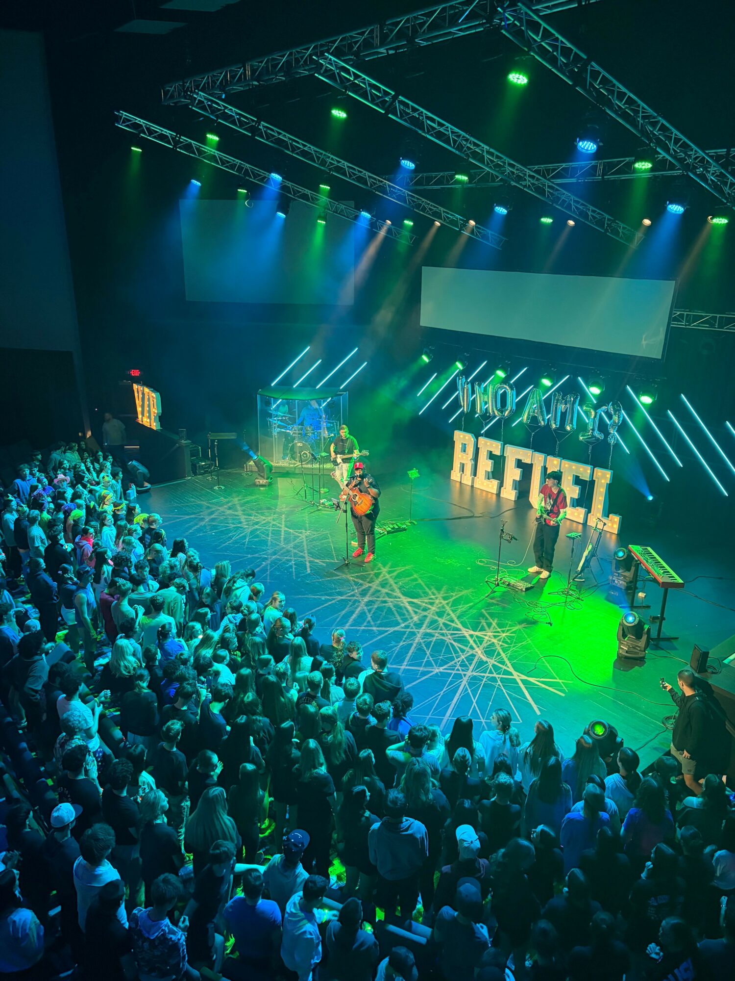 Crowd watching a stage with performers, colorful lights, and large screens at a concert or event.