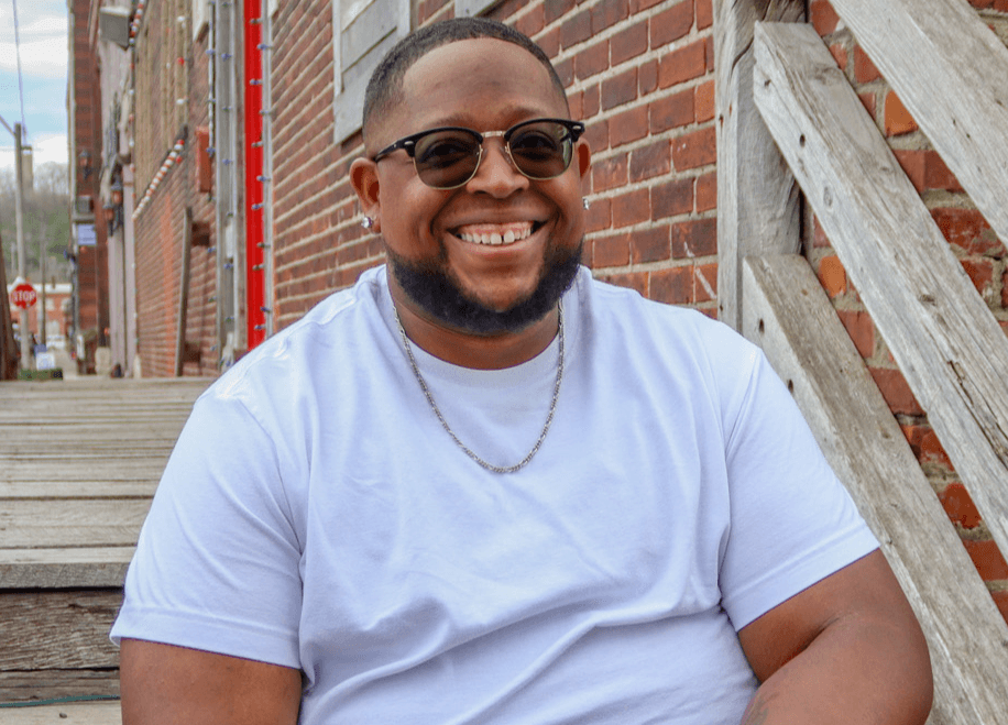 Smiling person with glasses and a chain, sitting outdoors on stairs, wearing a white t-shirt, brick wall background.