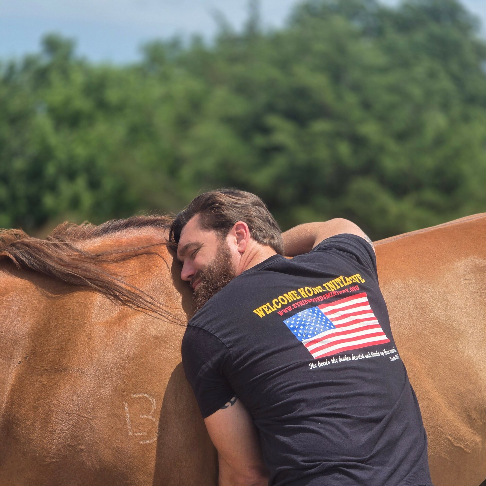 Man hugging a horse outdoors with green trees in the background.