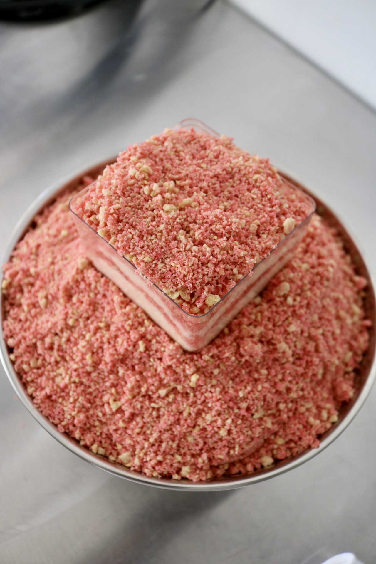 A pink layered cake with crumb topping in a round metal container, placed on a gray surface.