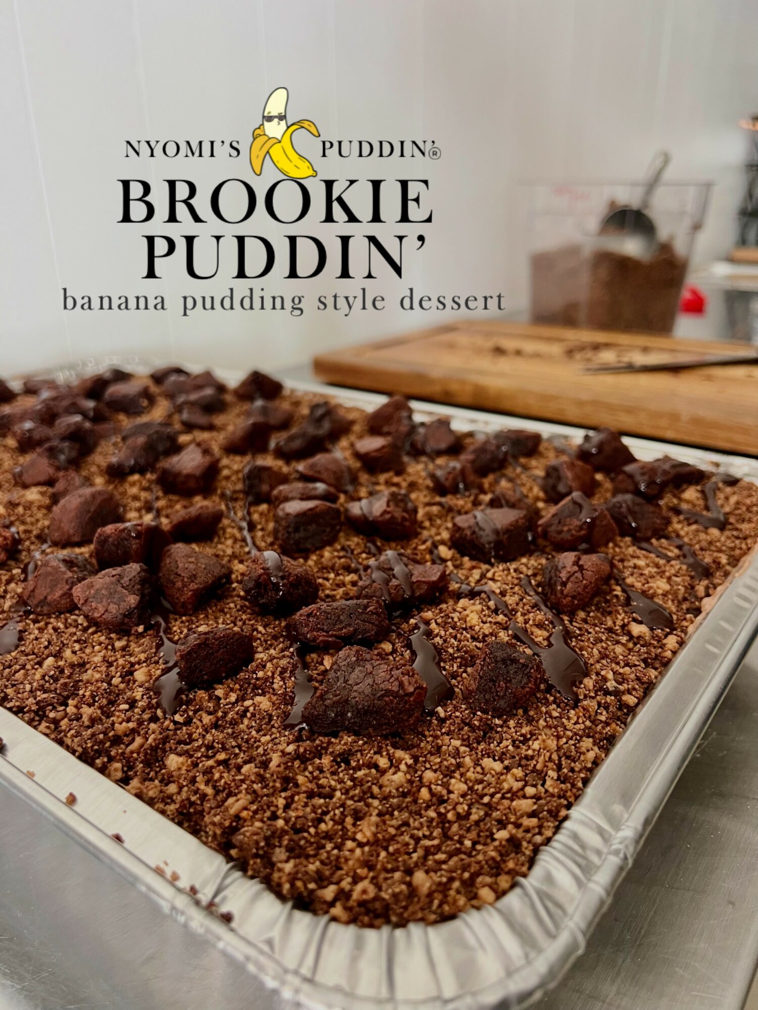 Banana pudding dessert with crumbled topping and chocolate chunks in a tray, with a wooden board and a jar in the background.